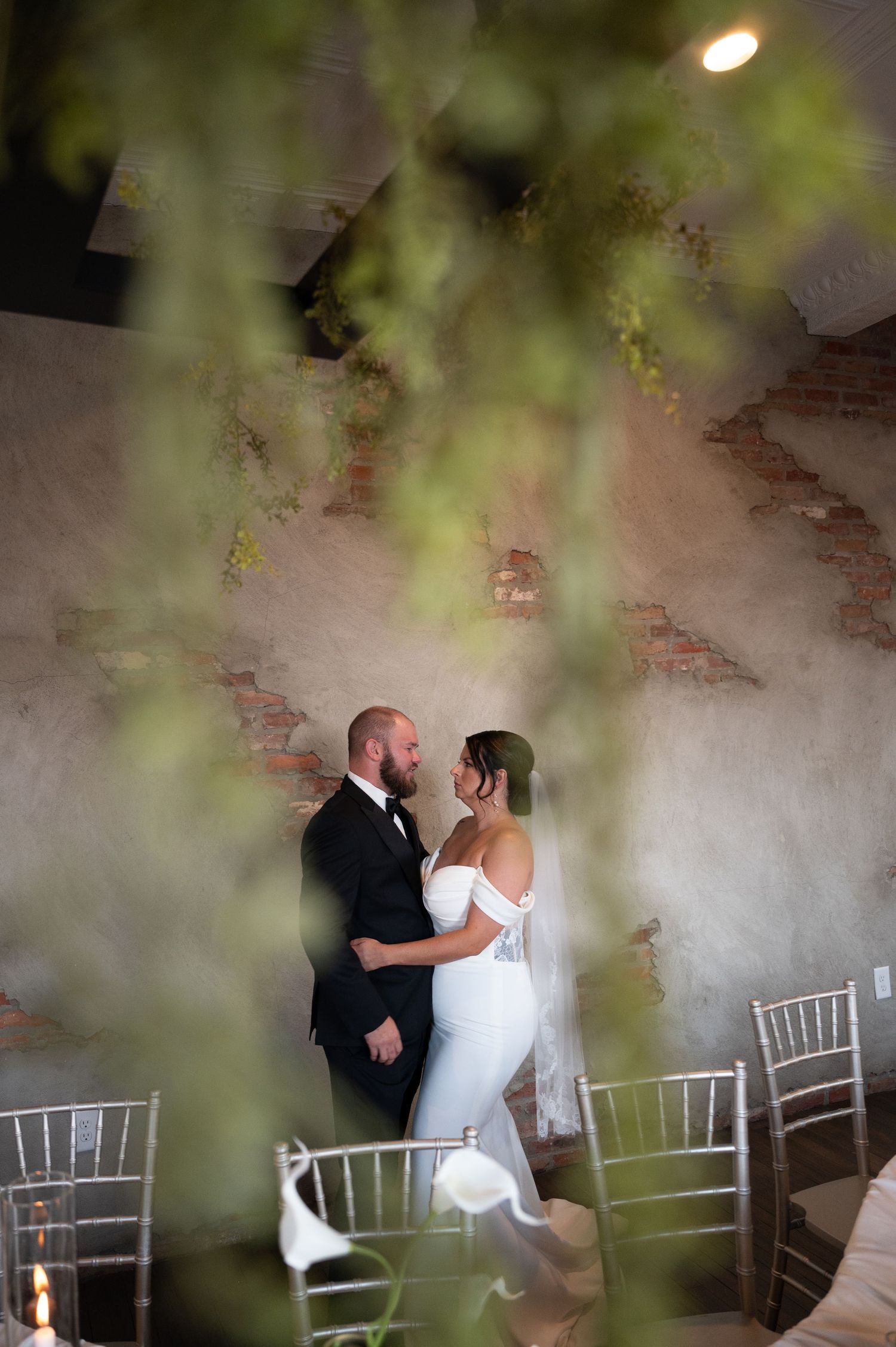 Bride and groom share a romantic moment in a rustic venue with exposed brick walls and hanging greenery.