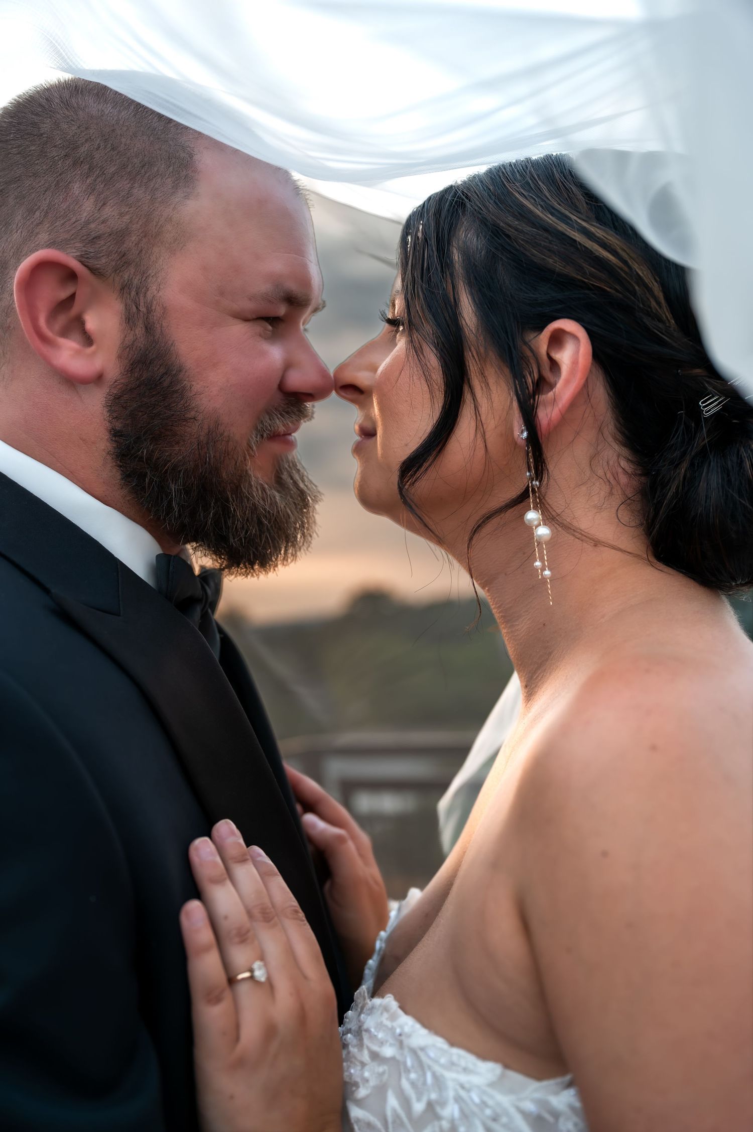 Bride and groom share an intimate moment under a wedding veil, nearly touching noses, bride wearing pearl earrings and diamond ring.