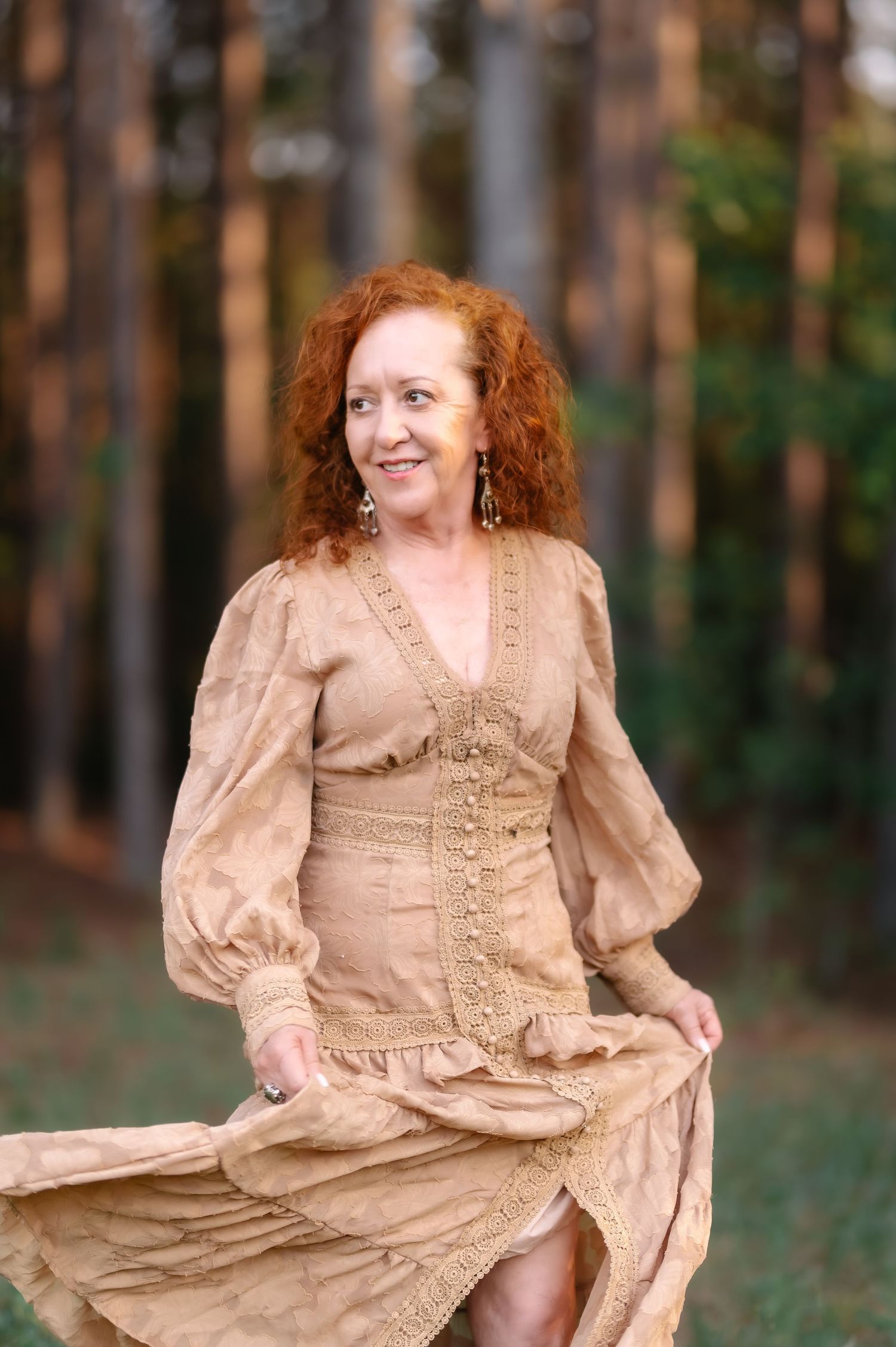 A smiling woman with curly red hair wearing a golden boho dress twirls outdoors in a pine forest setting.