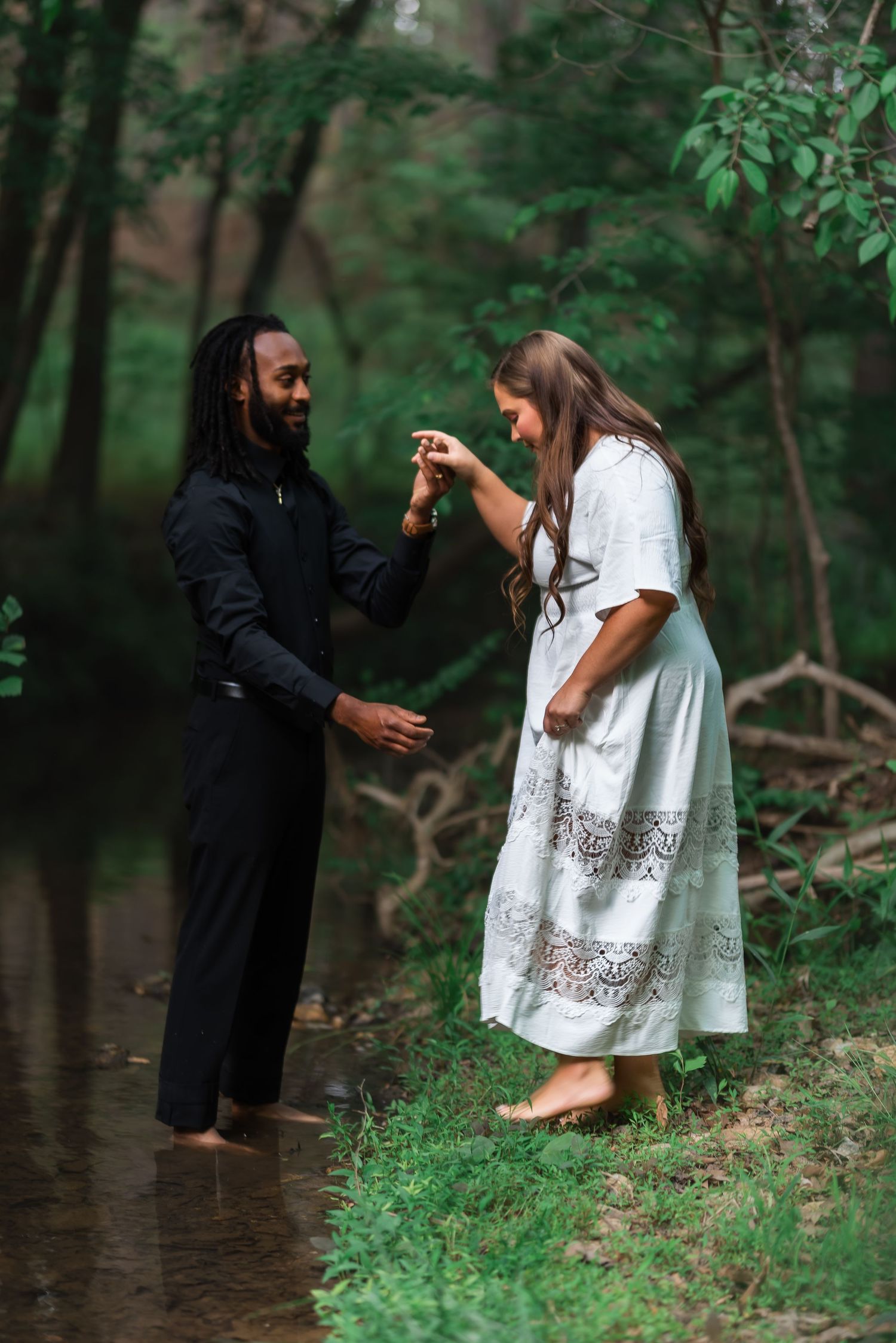 Couple holds hands near a forest creek, man in black outfit, woman in white lace dress.