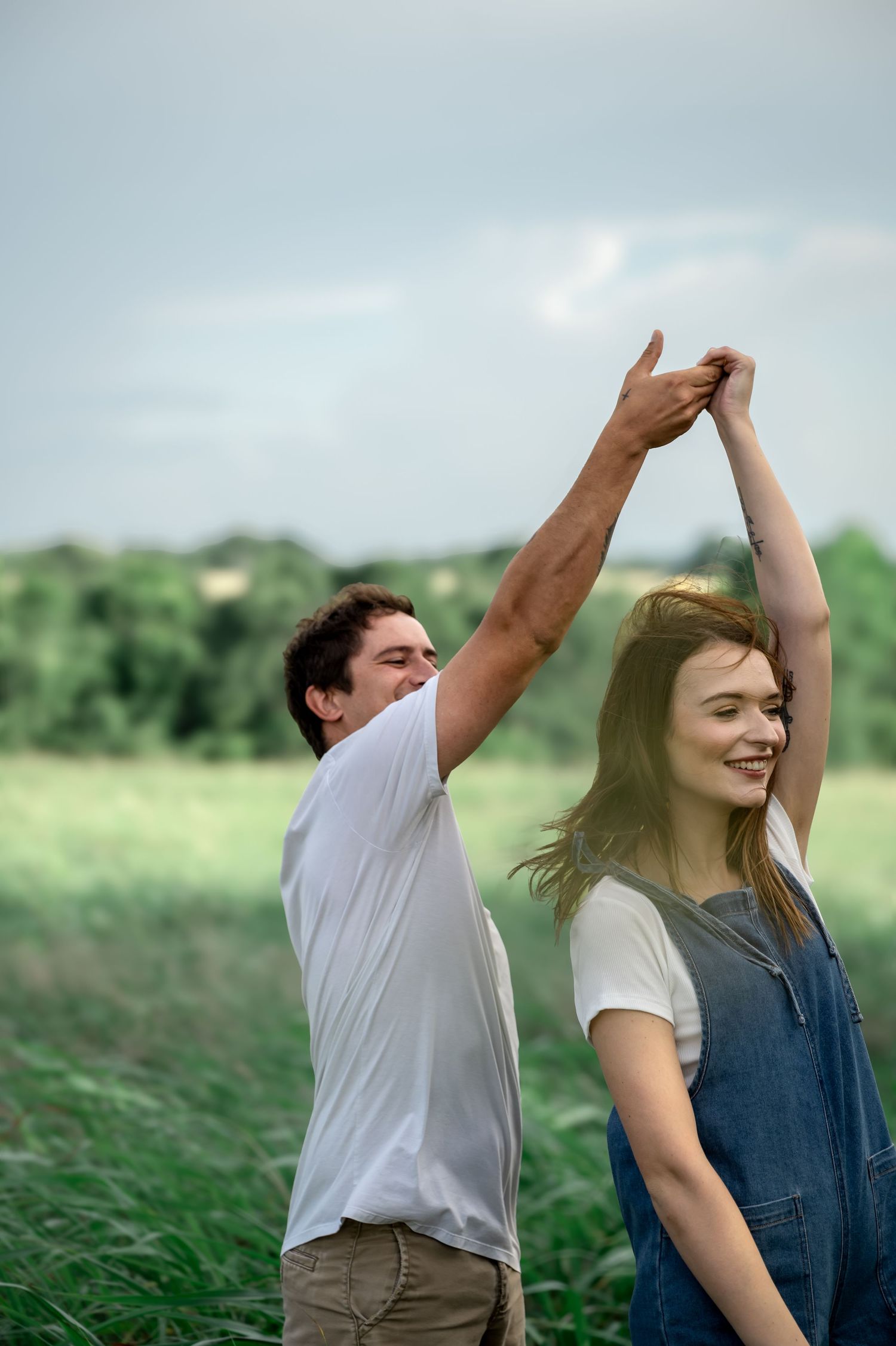 Couple dancing in green field, man spinning woman with raised hands, smiling outdoors on cloudy day.