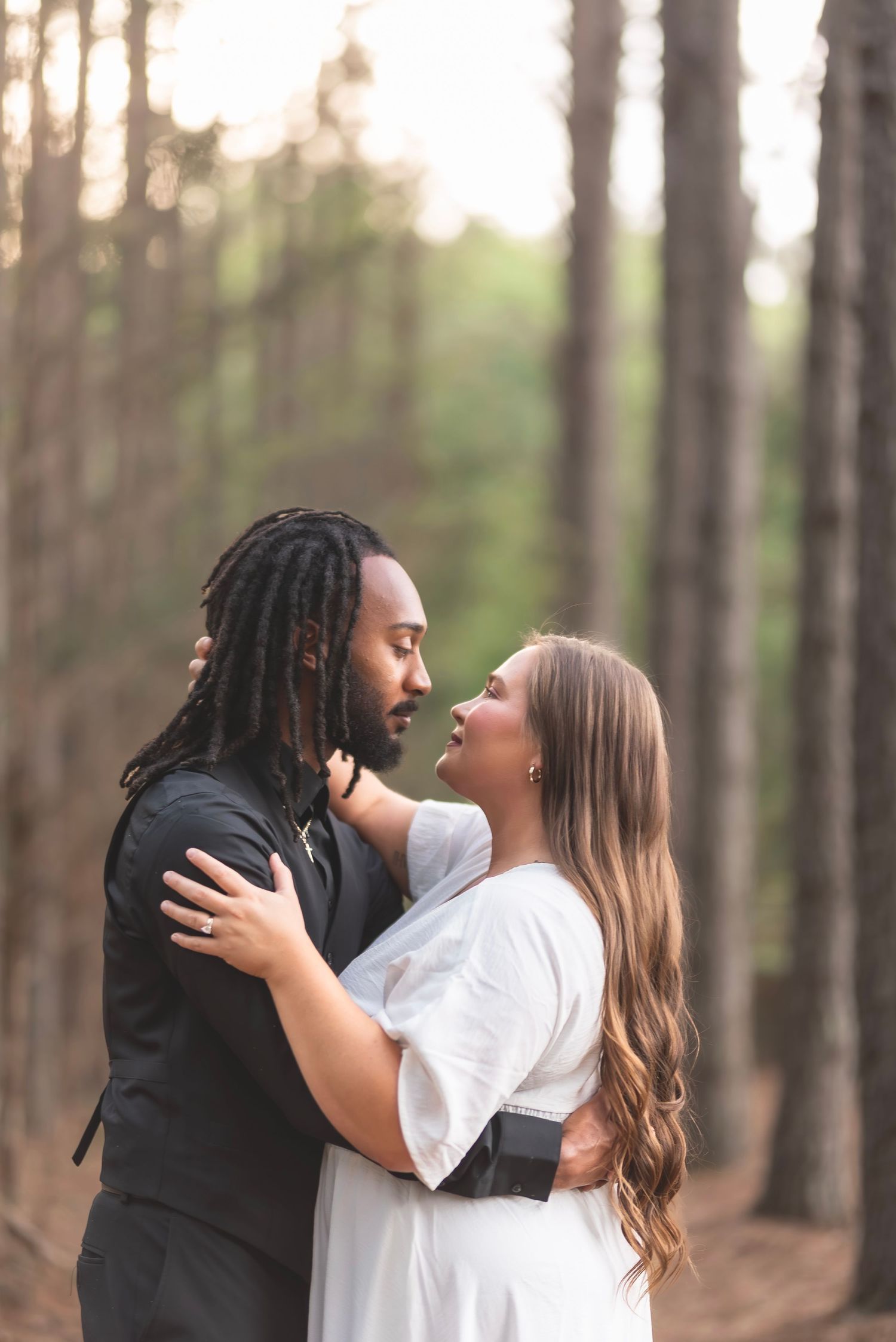 A couple embracing and gazing at each other romantically among tall pine trees at sunset.