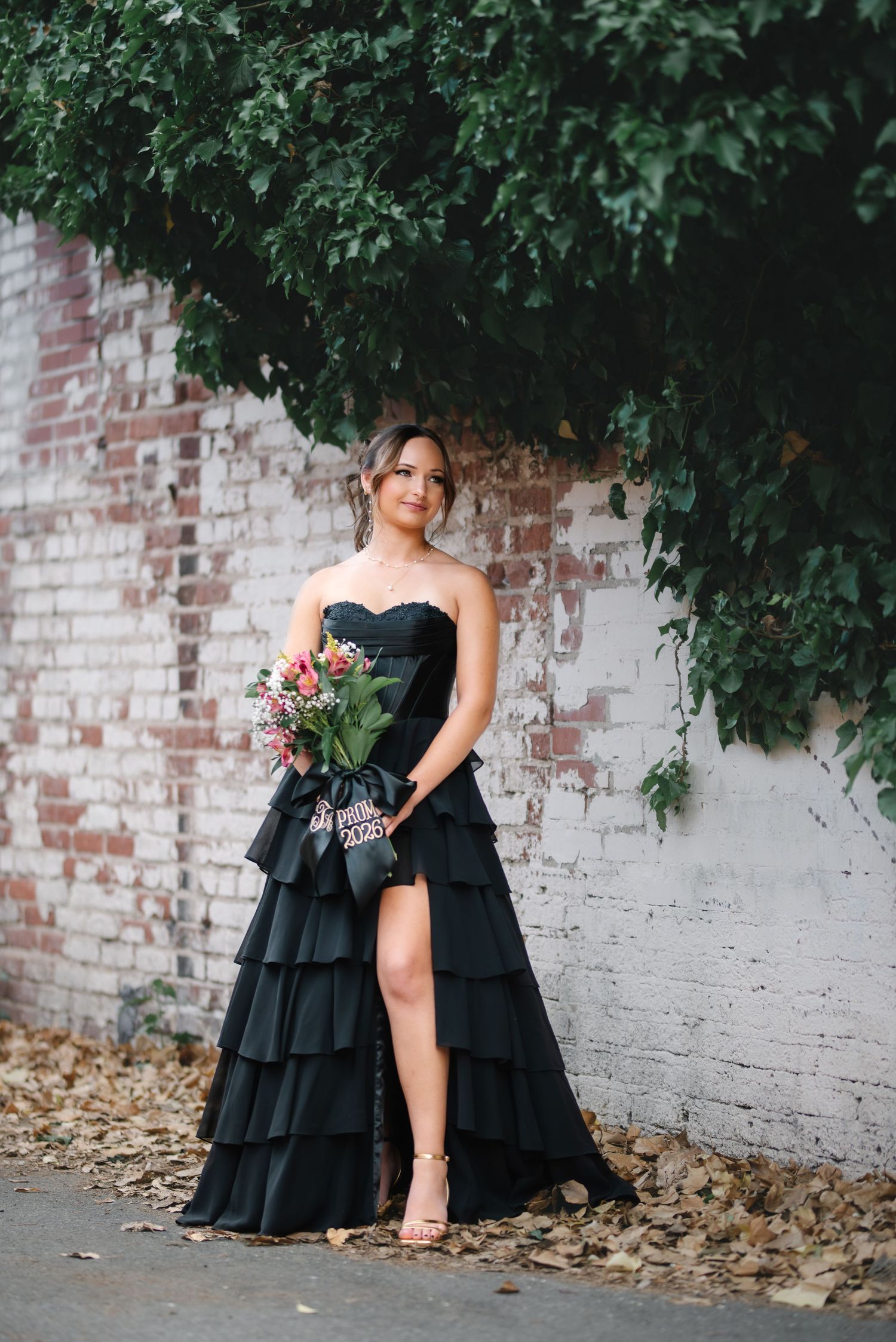 A woman in a black strapless tiered gown holds a colorful bouquet near a brick wall covered in ivy.