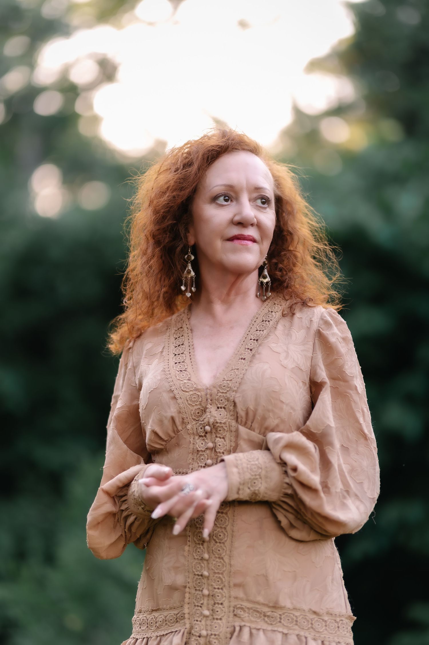Woman with curly red hair wearing a tan lace dress poses outdoors with green trees in the background.