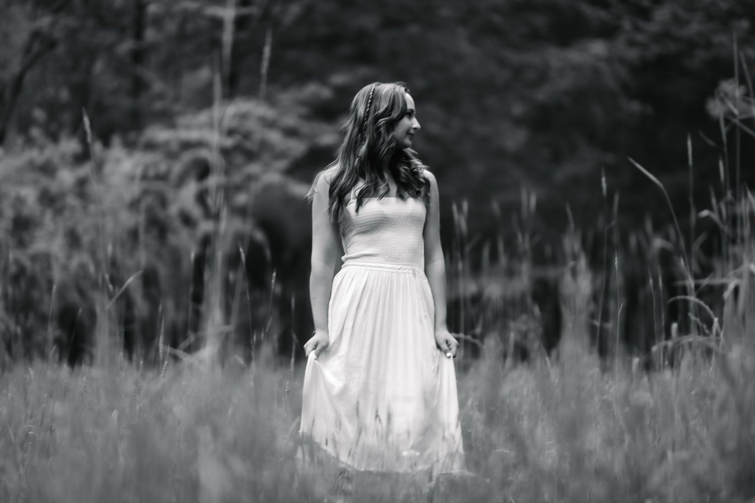 Black and white photo of a woman in a white dress standing in a tall grass field surrounded by trees.