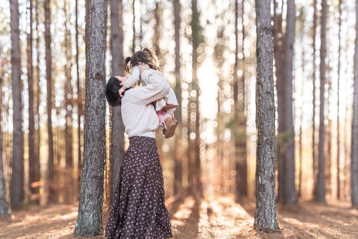 Mother lifting child in sunlit pine forest, sharing a joyful moment in warm autumn light.