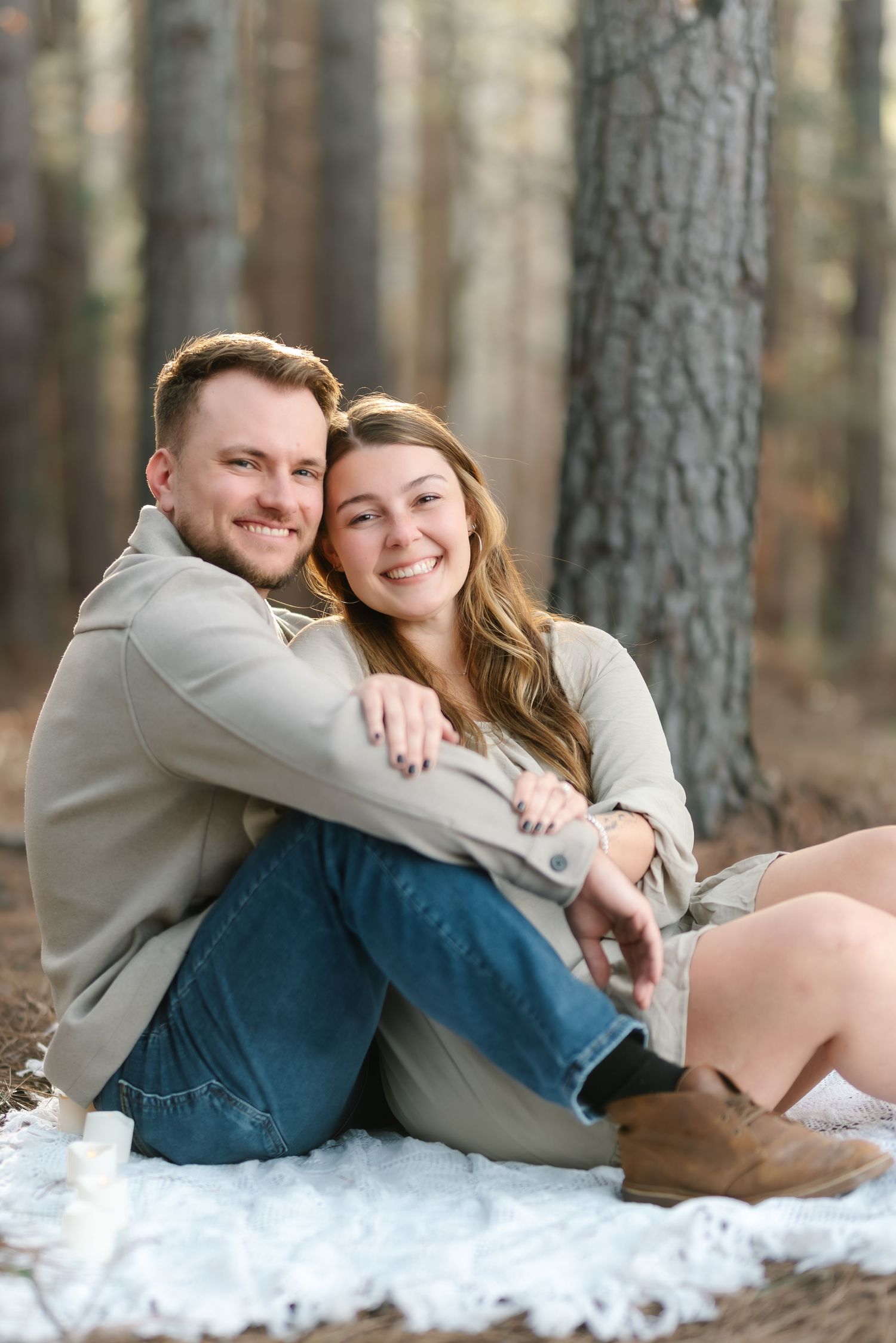 A smiling couple sits together on a blanket in a pine forest, embracing warmly in casual neutral and denim outfits.