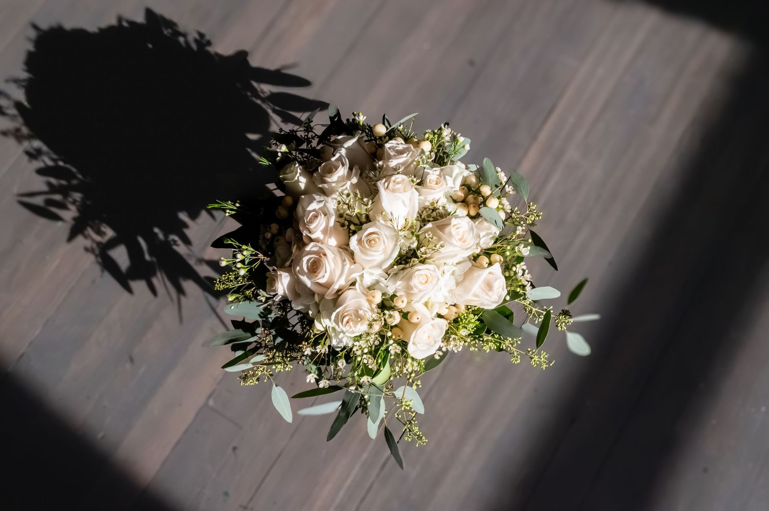 Elegant bridal bouquet of white roses and greenery on dark wood floor with soft shadows.