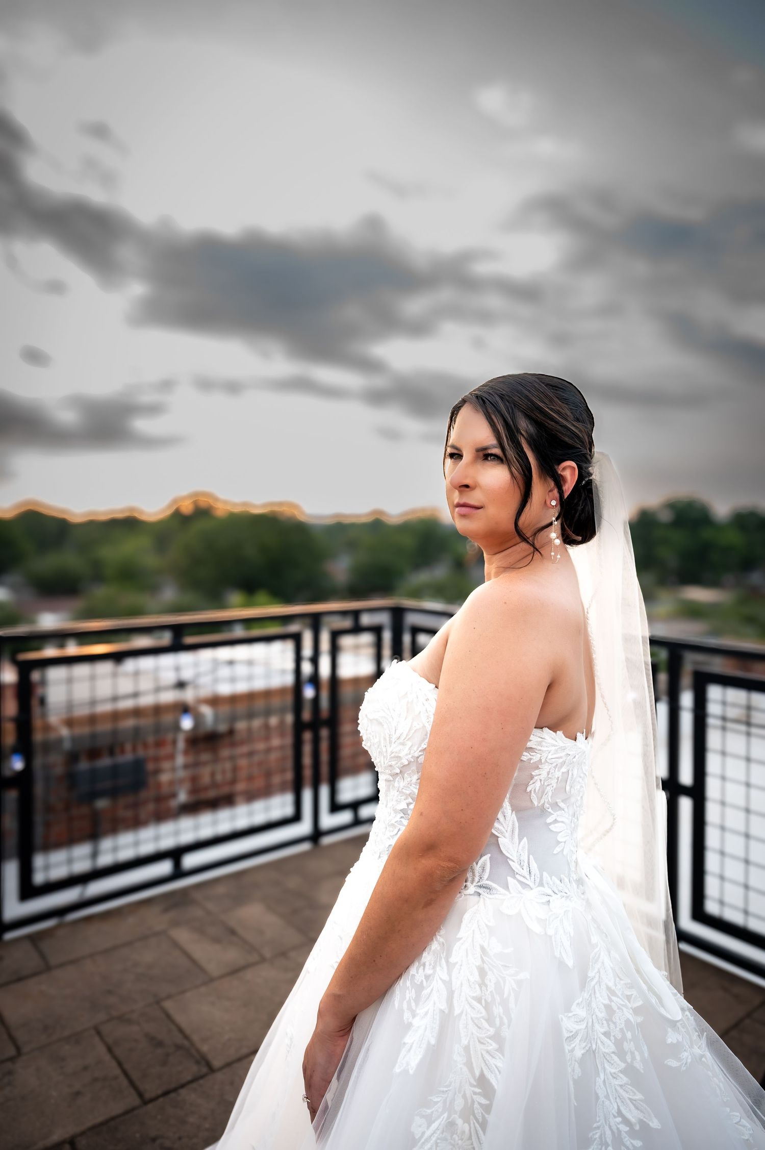 Bride in strapless lace wedding gown and veil poses on rooftop terrace against cloudy sky.