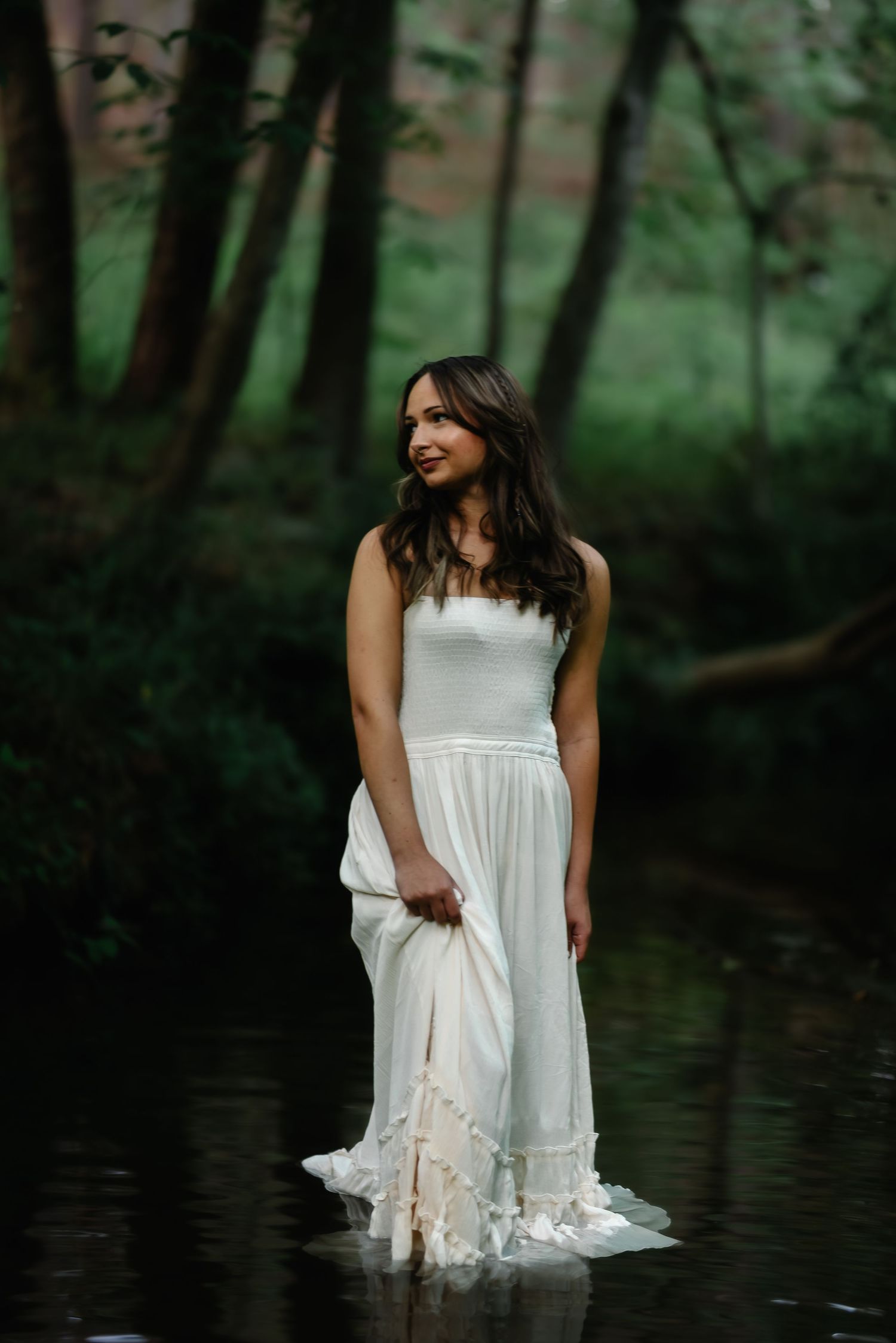 Woman in white dress standing in forest stream, surrounded by lush green trees in a dreamy, ethereal outdoor setting.