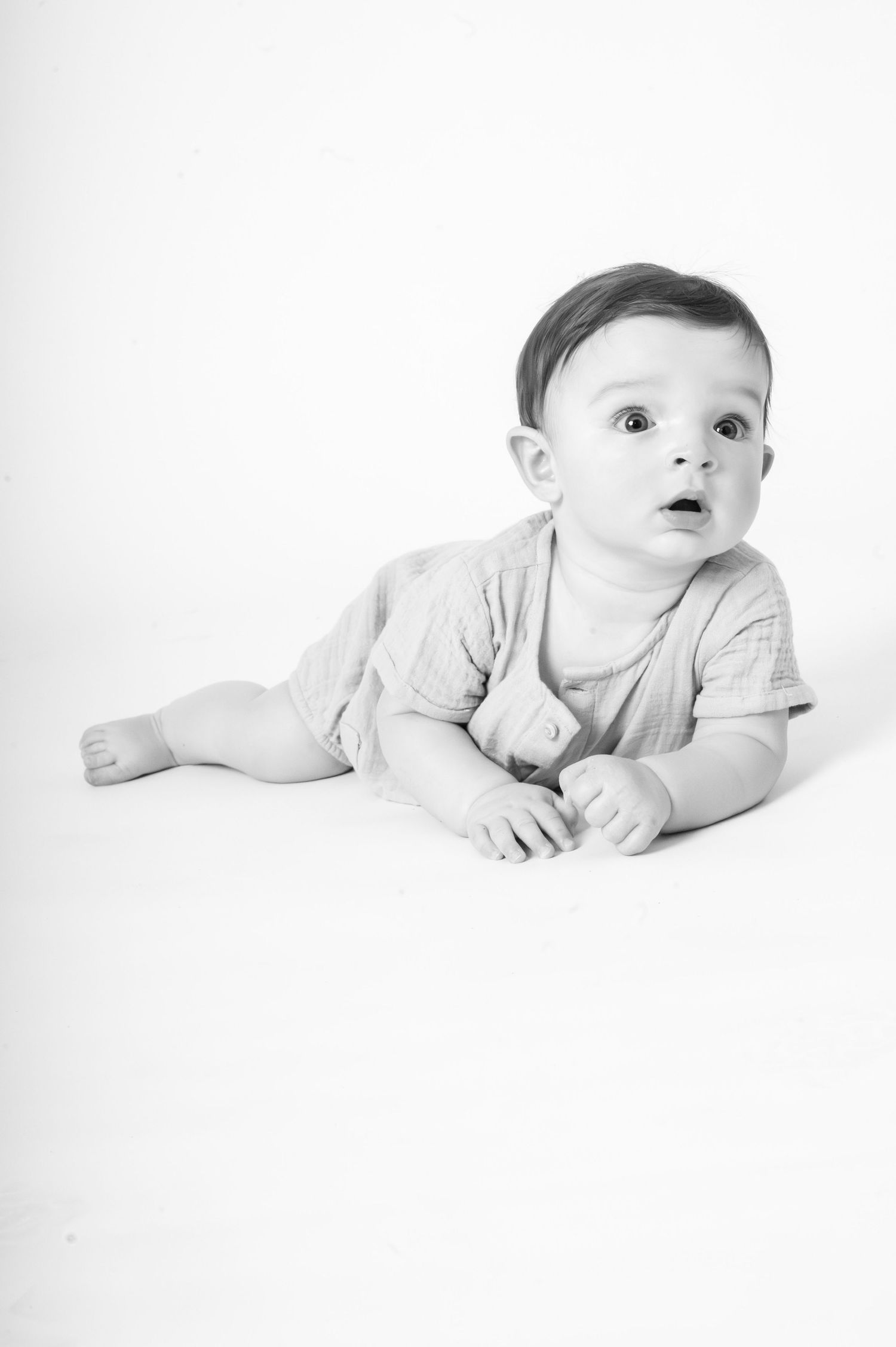 Black and white photo of a curious baby lying on white background wearing a light romper.