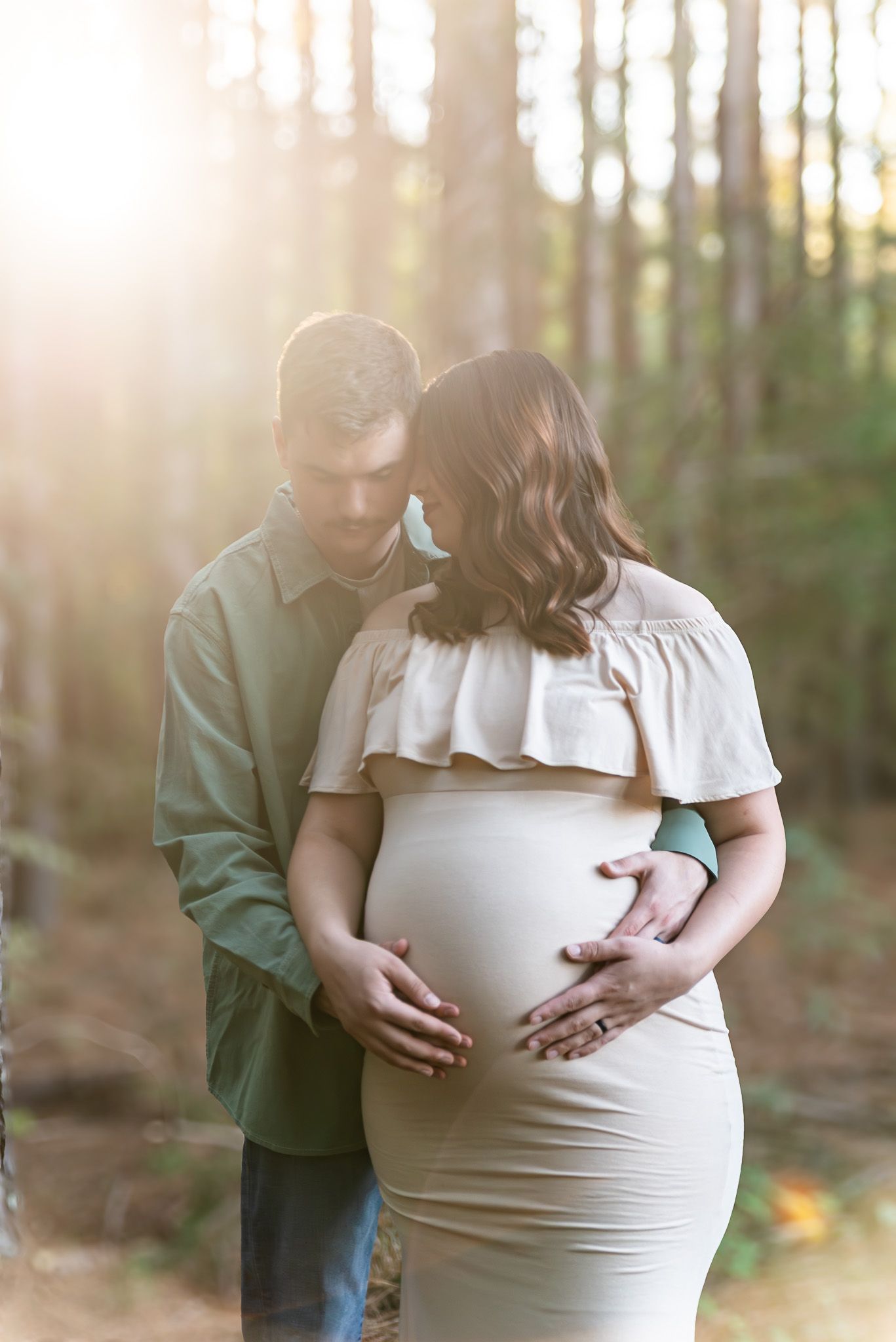 Couple tenderly cradling pregnant belly in sunlit pine forest maternity portrait.