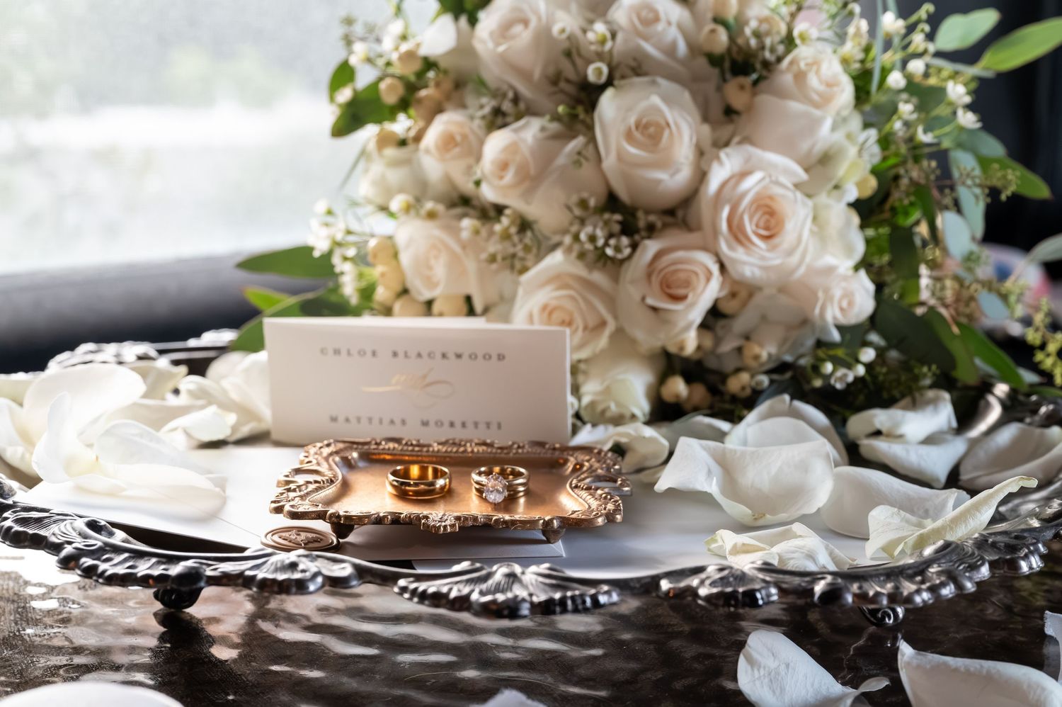 Elegant wedding flat lay with white roses bouquet, gold rings, and place card on ornate silver tray.