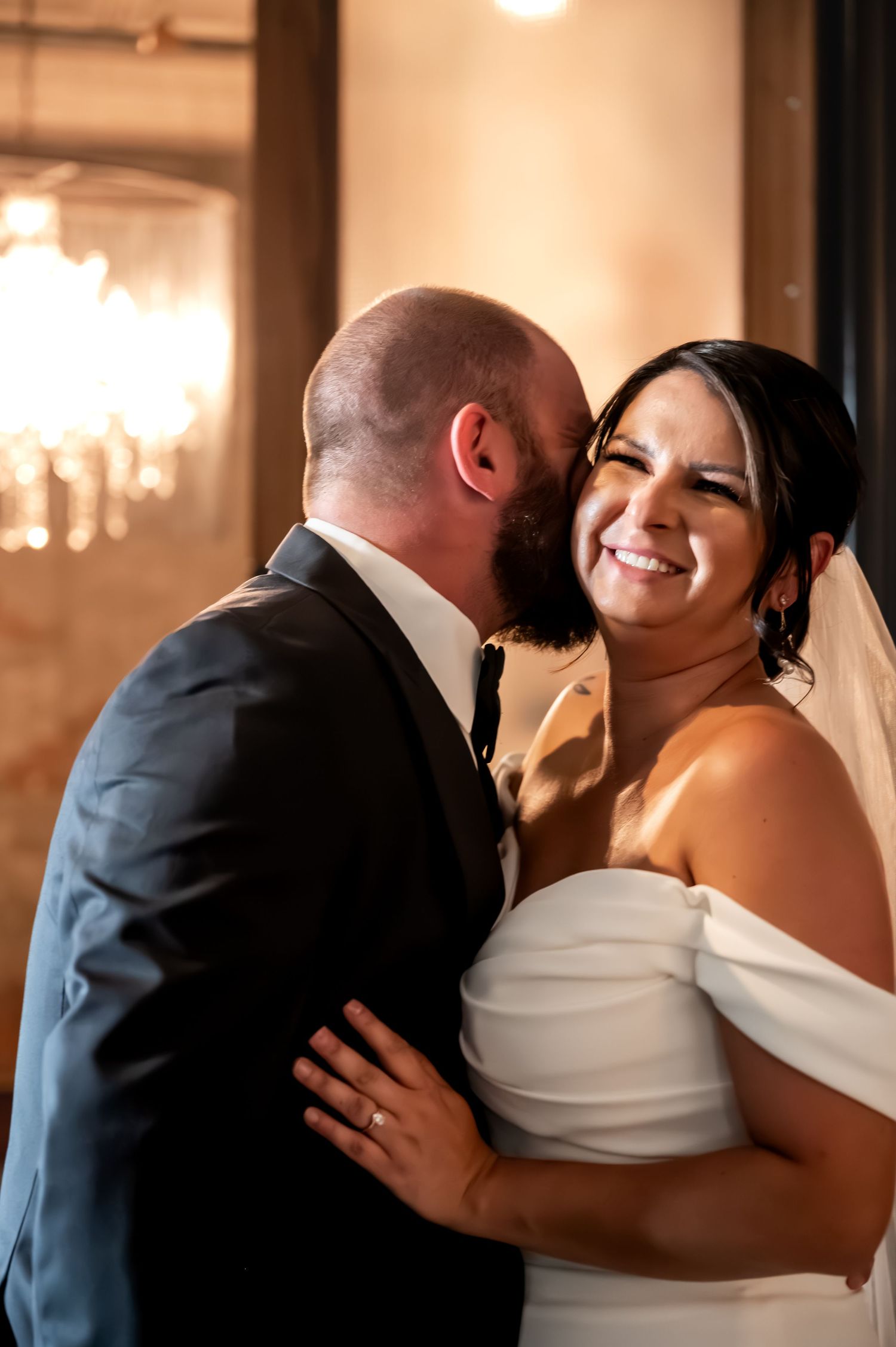 Groom kisses bride on cheek as she smiles joyfully, wearing off-shoulder white gown and veil in elegant venue.