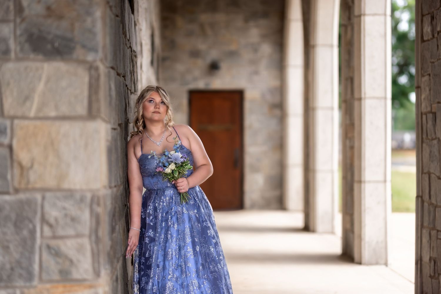 Young woman in blue floral gown holding flowers, leaning against stone wall in elegant outdoor corridor.