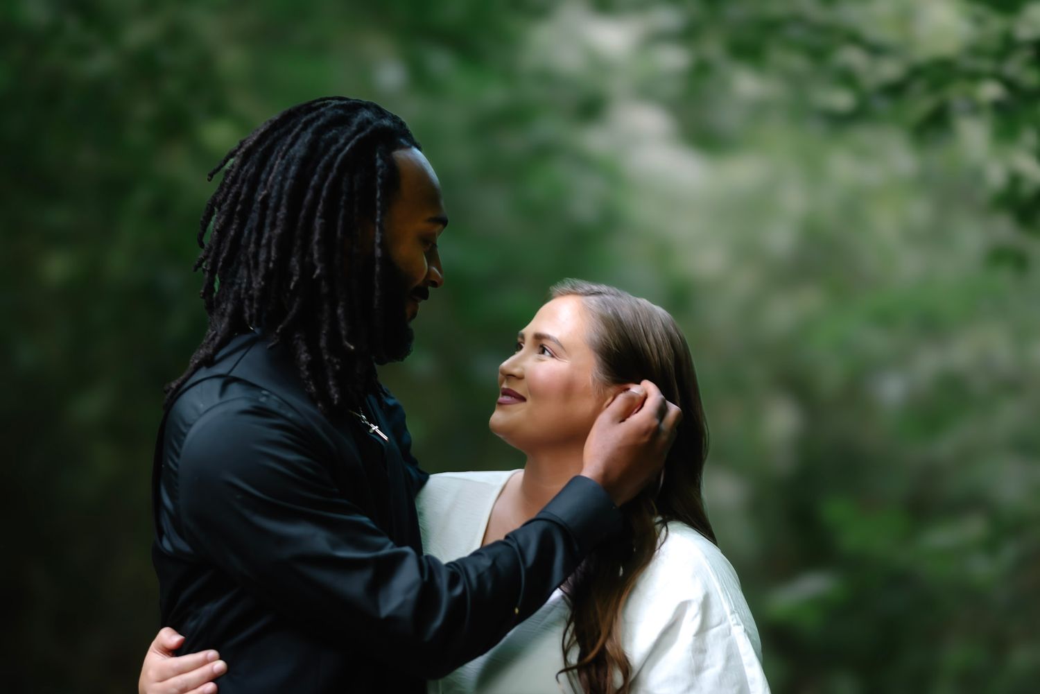 A couple shares a tender moment outdoors, with a man gently touching a woman's hair amid lush green trees.