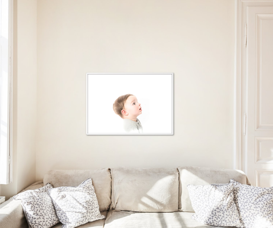 Framed portrait print of a child blowing a kiss hung above a neutral beige sofa with patterned cushions in a bright room.