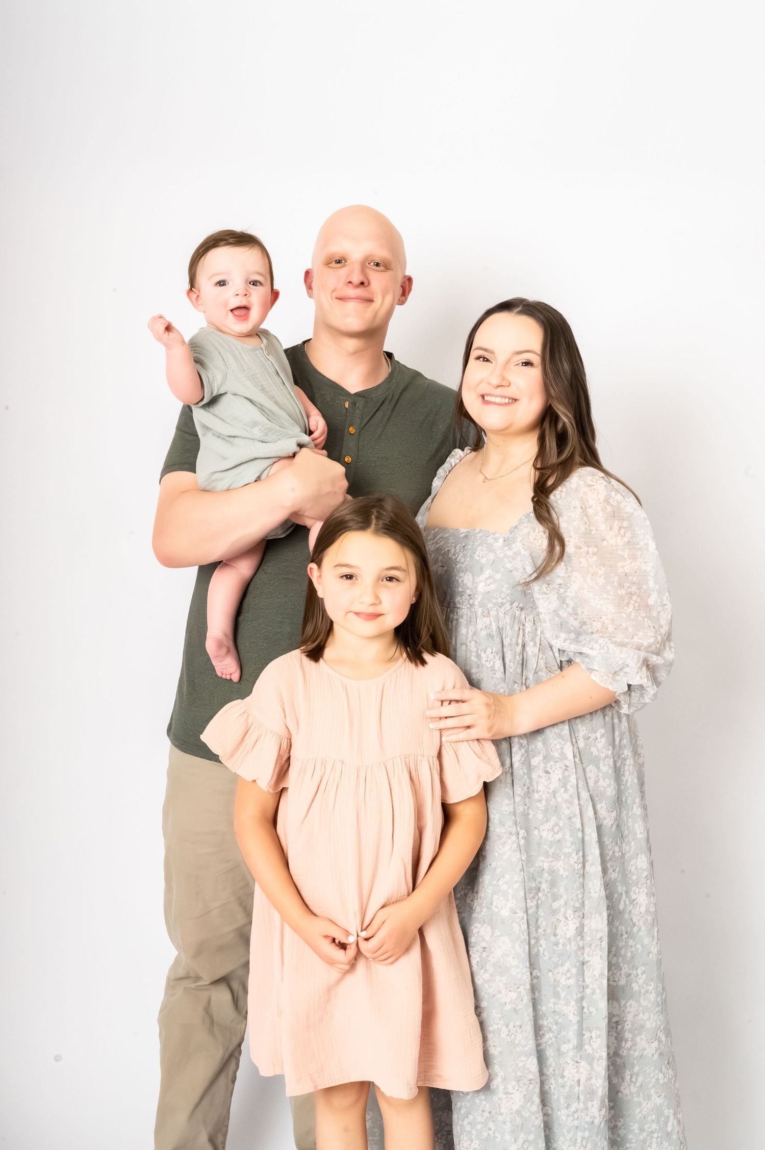 A happy family of four posing together, with dad holding a baby, mom in a floral dress, and a young girl in pink.