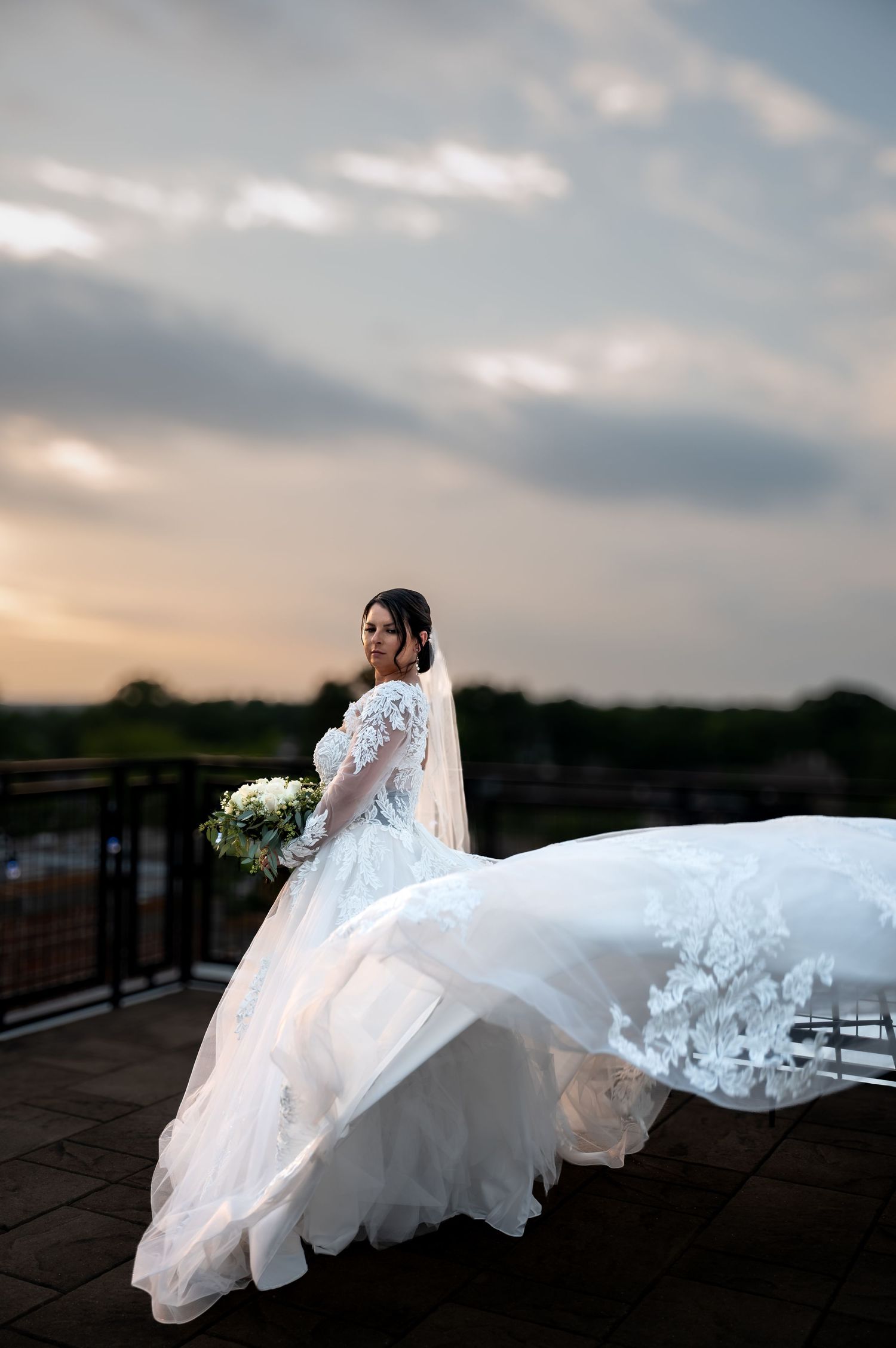 Bride in flowing white lace gown with veil billowing in wind holds bouquet on rooftop at sunset.