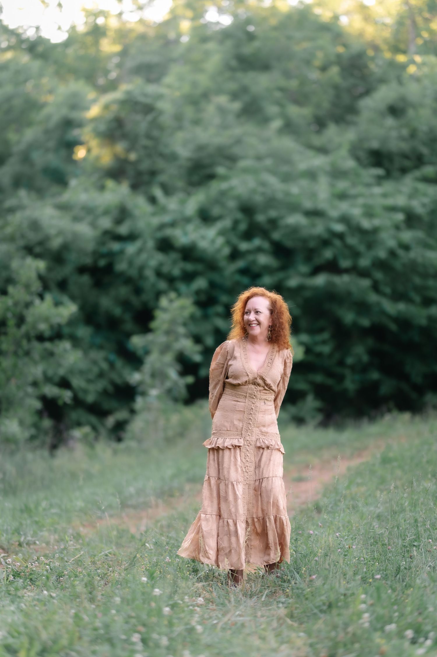 A smiling redhead woman in a flowing beige dress walks along a grassy path surrounded by lush green trees.