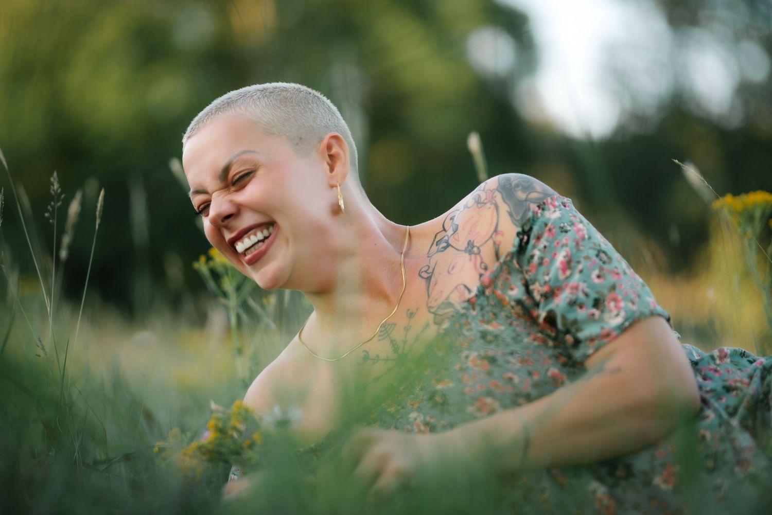 A joyful woman with a shaved head laughs while sitting in a lush green field, wearing a floral dress.