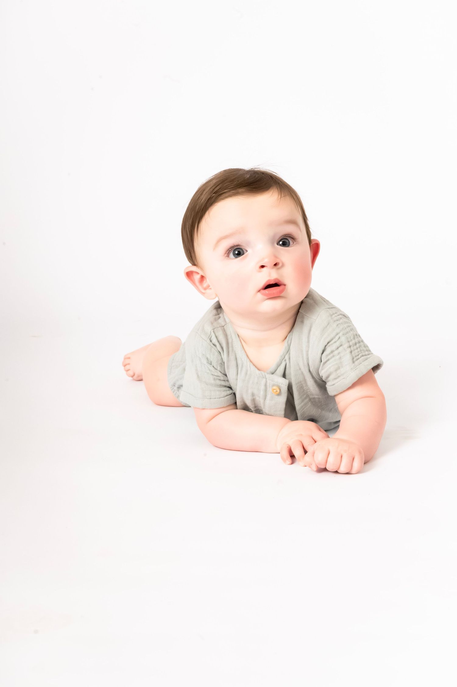 Adorable baby in a gray romper lying on white background, looking up with curious wide eyes.