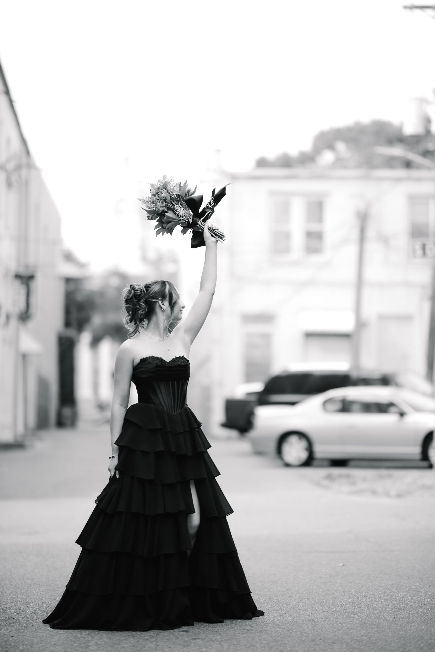 Bride in black strapless ruffled gown raises bouquet triumphantly on urban street in black and white photo.
