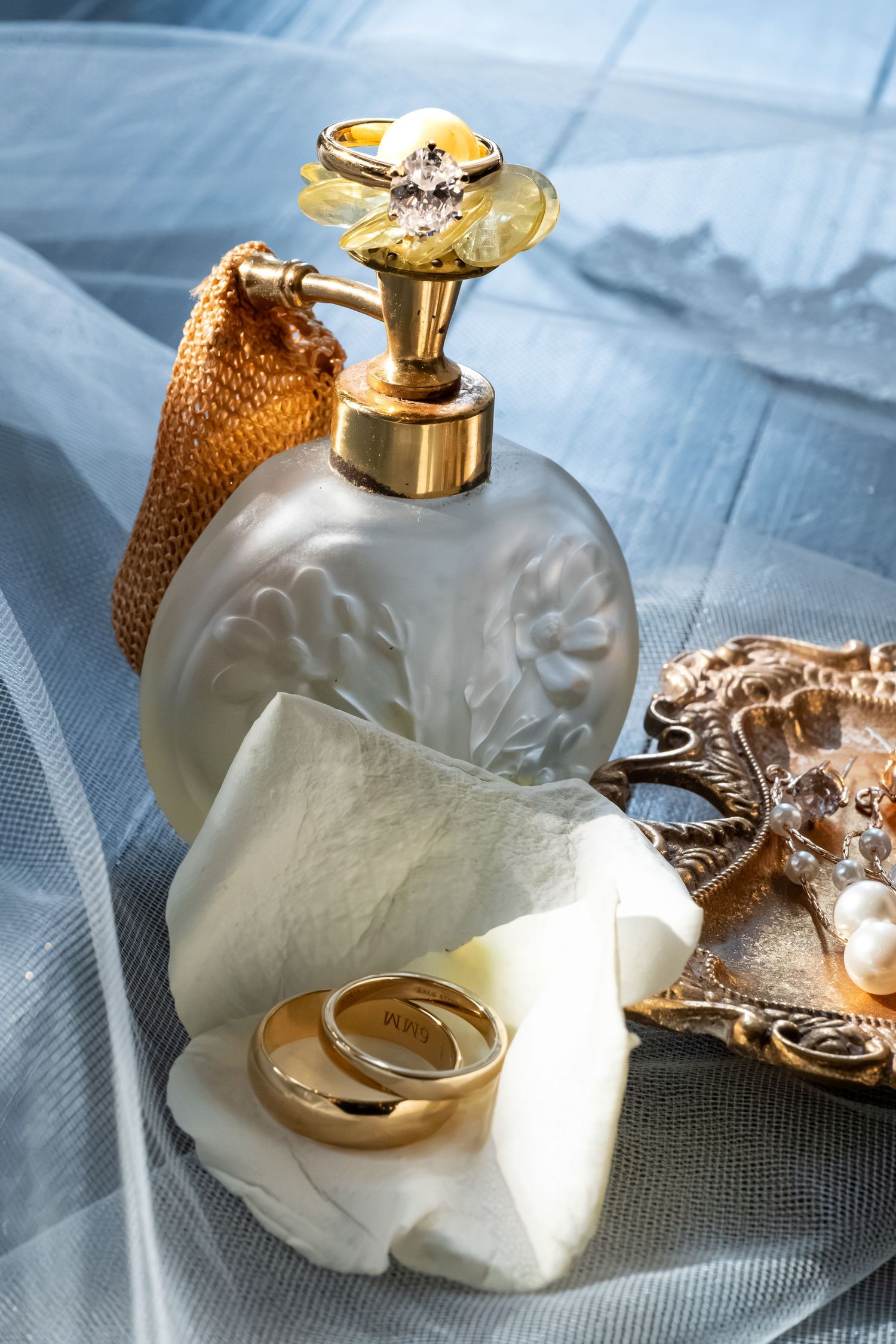 Elegant perfume bottle with gold rings, pearl jewelry, and sheer blue fabric for a luxurious bridal vanity display.