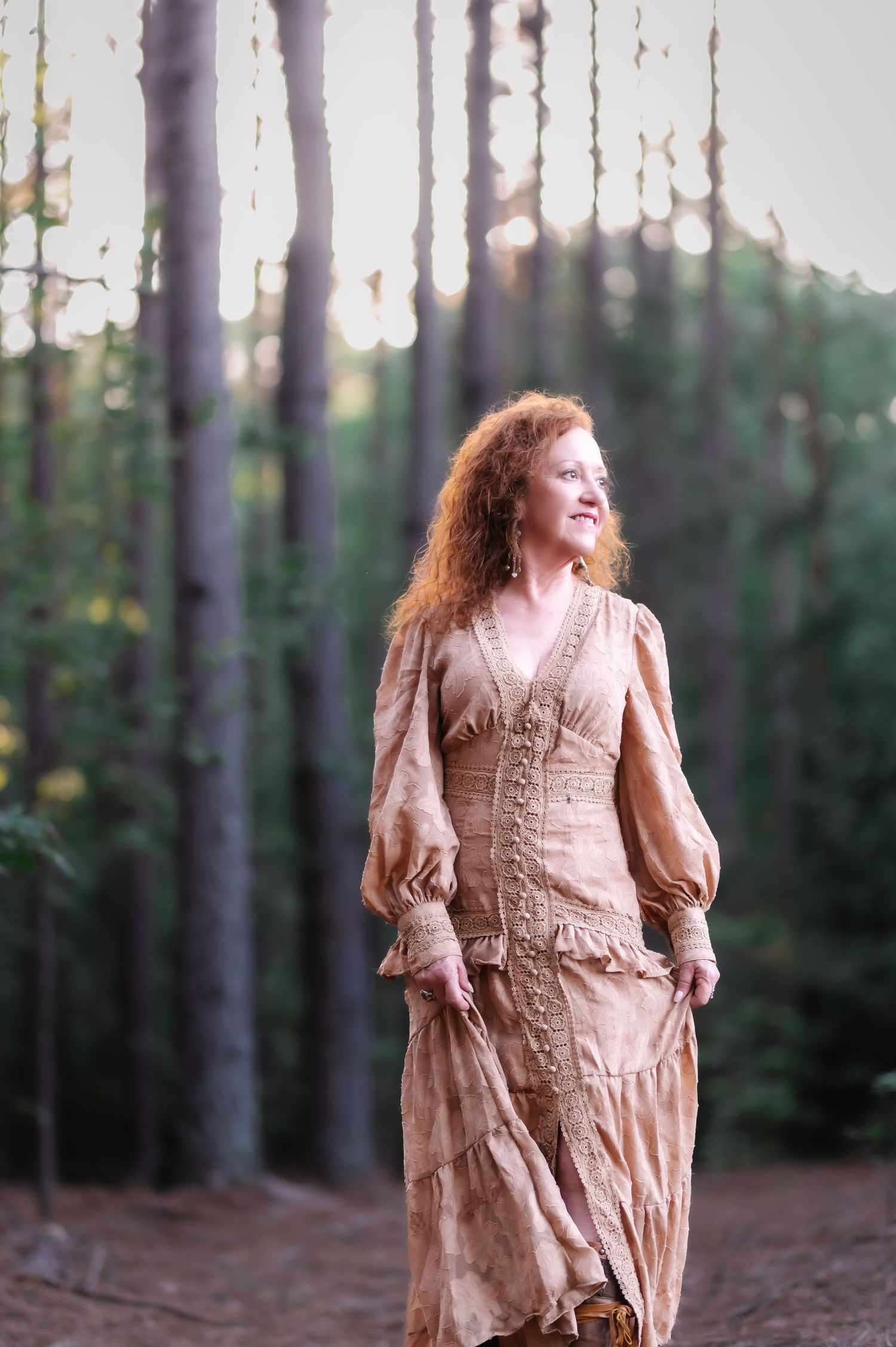 Red-haired woman in vintage beige dress standing gracefully among tall trees in a sunlit forest.