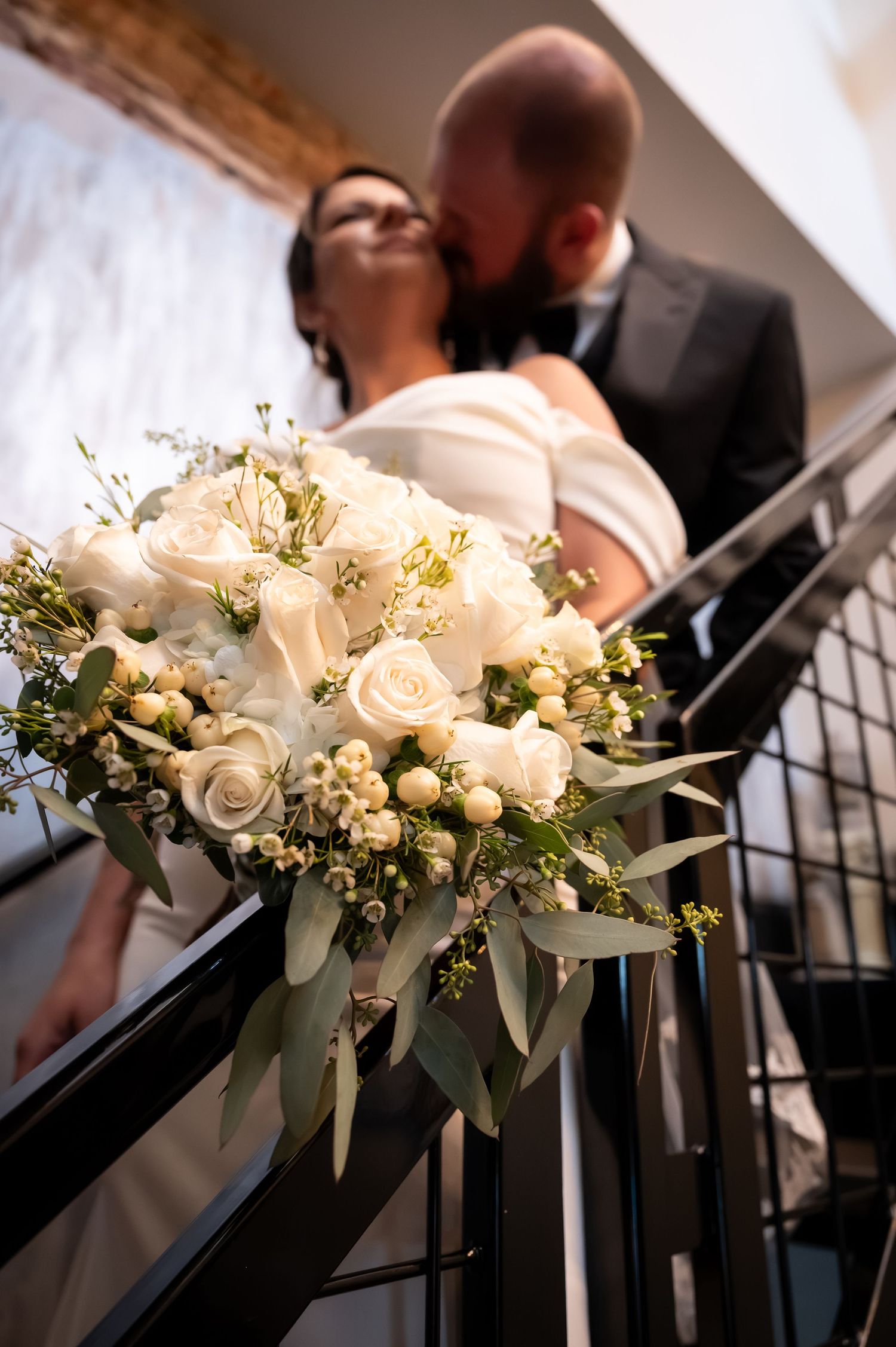 A wedding couple shares a kiss on a staircase, with a lush white rose bridal bouquet in the foreground.