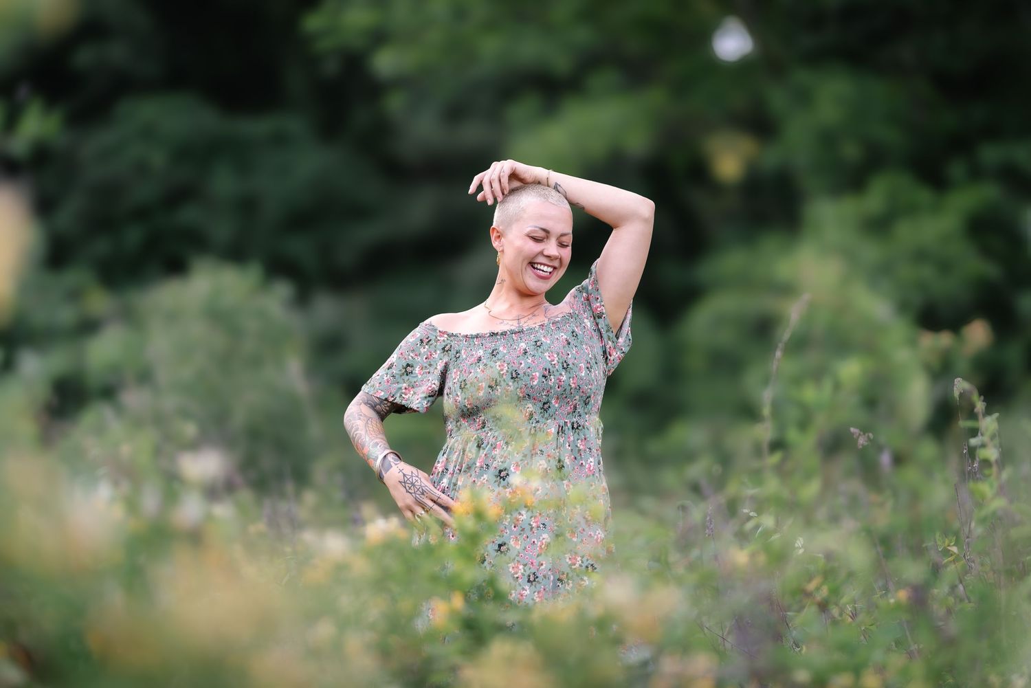 Laughing woman with tattoos in floral dress posing playfully in a wildflower meadow.