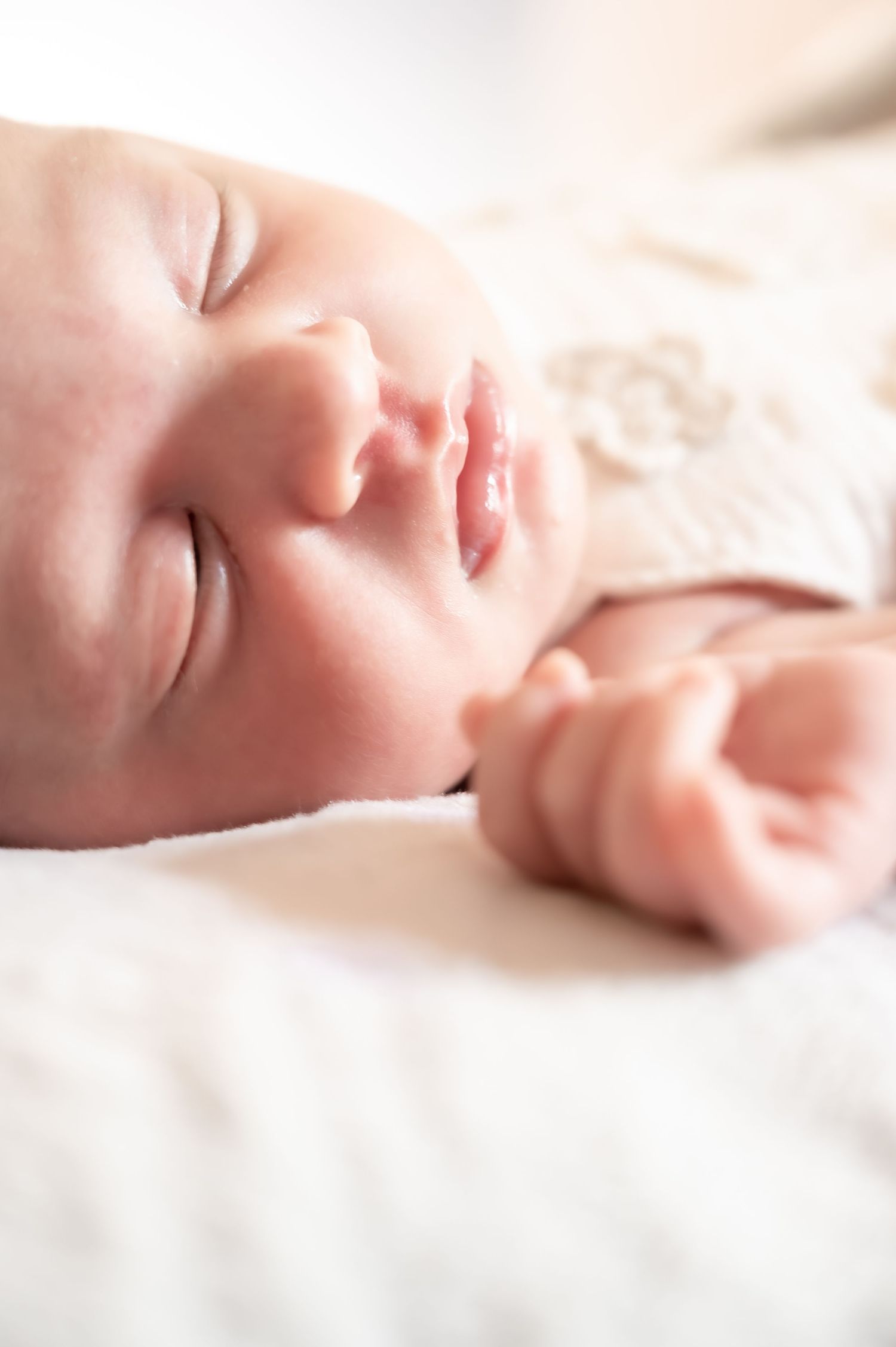 A peaceful newborn baby sleeping on a soft white blanket, wearing a delicate floral outfit, with tiny hands visible.