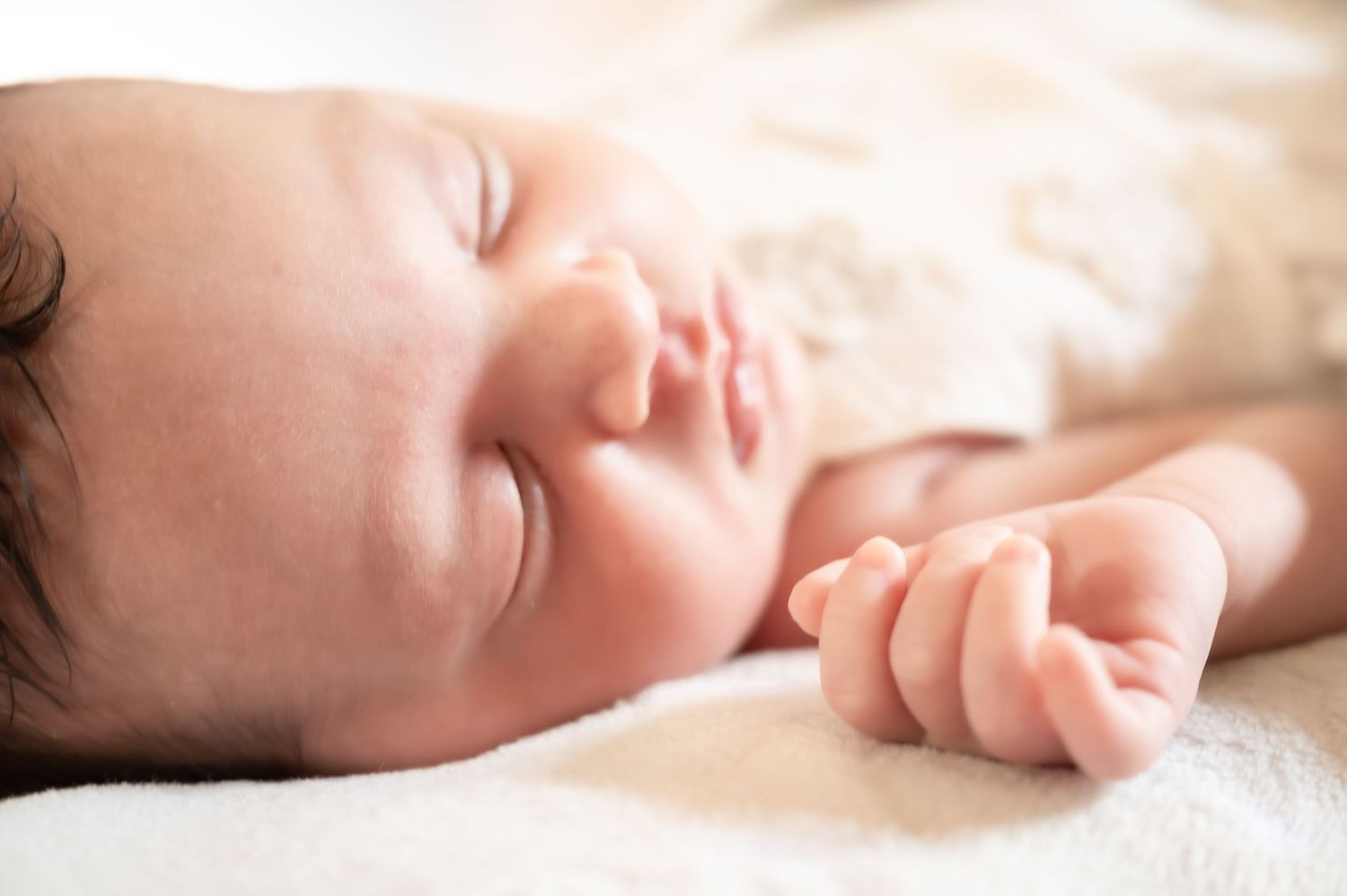 Peaceful newborn baby sleeping on a soft white blanket with tiny curled fingers and dark hair.