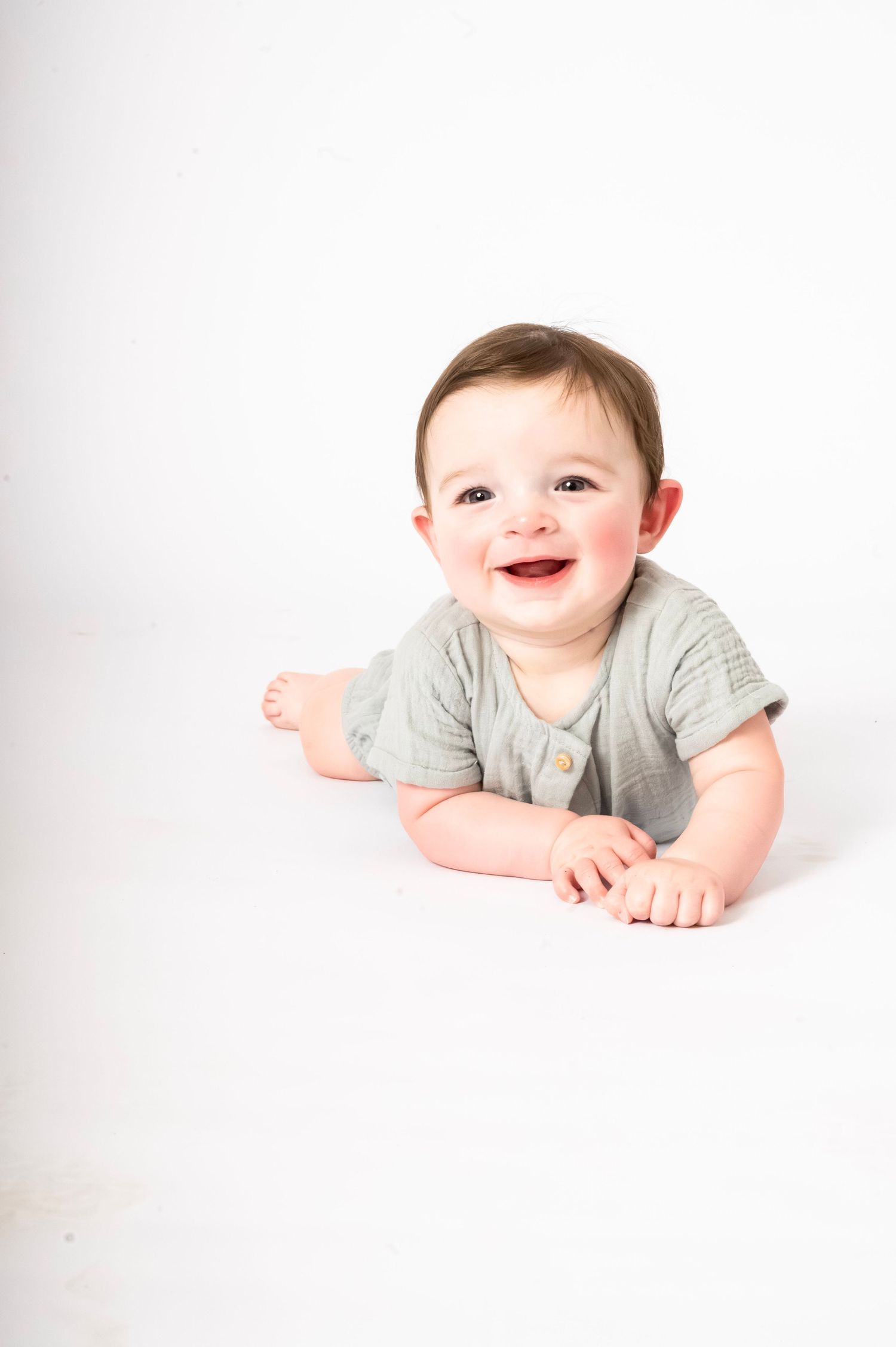 Smiling baby in a sage green romper lying on a white background, looking up with bright eyes and a joyful expression.