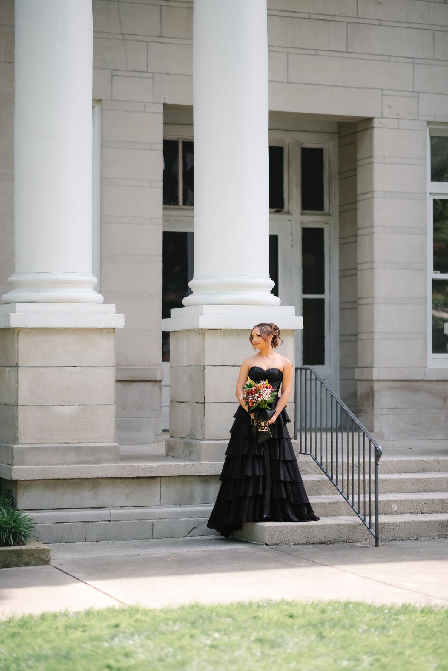 Bride in black strapless gown holding colorful bouquet stands before white stone building with tall columns.