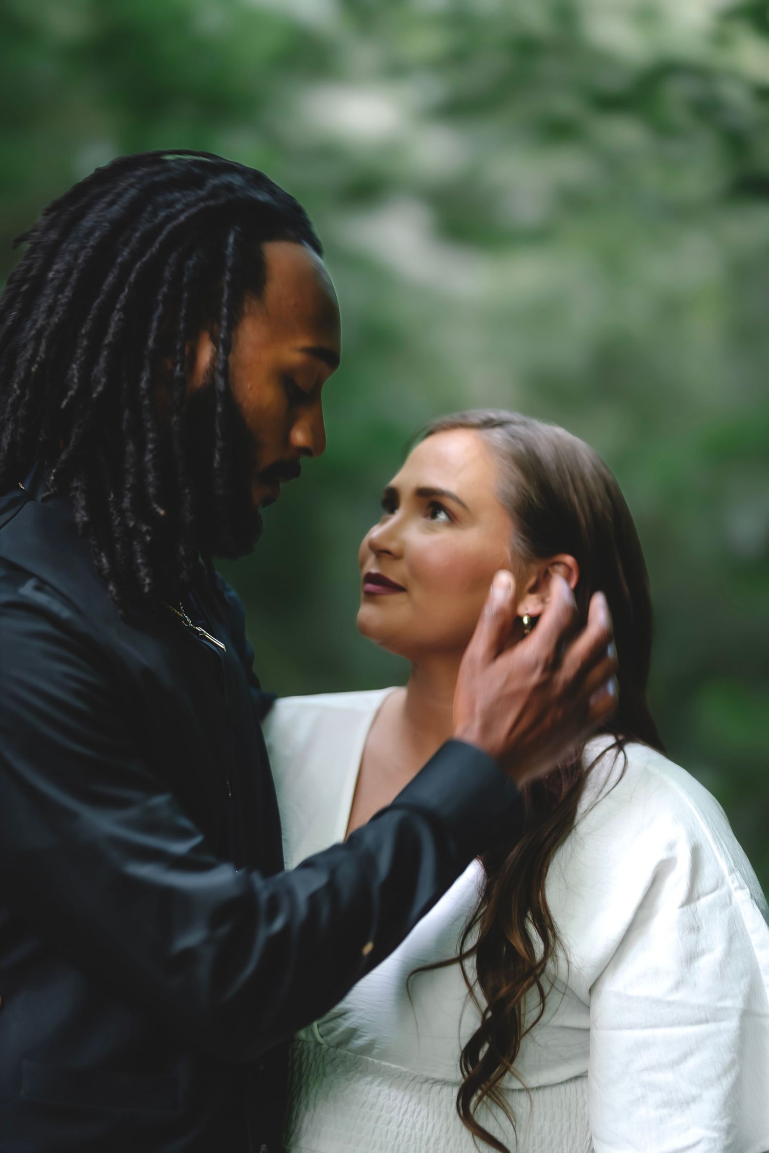 A tender moment between a couple in a lush green forest, the man gently cradling the womans face.