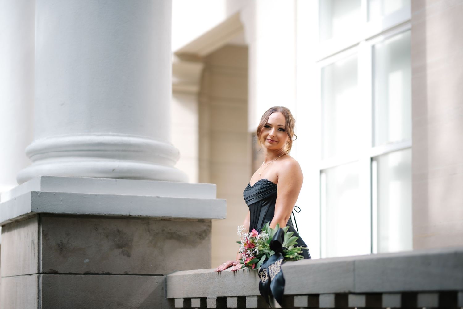 Bride in black strapless gown sitting on stone ledge with bouquet beside white columns.