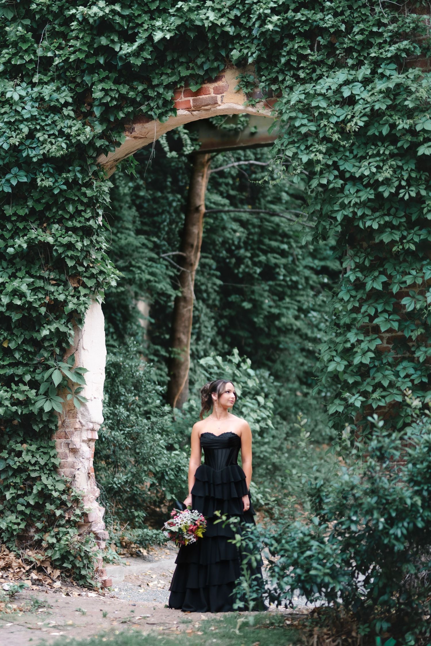 Woman in black strapless gown holding bouquet stands before ivy-covered brick archway in lush garden setting.