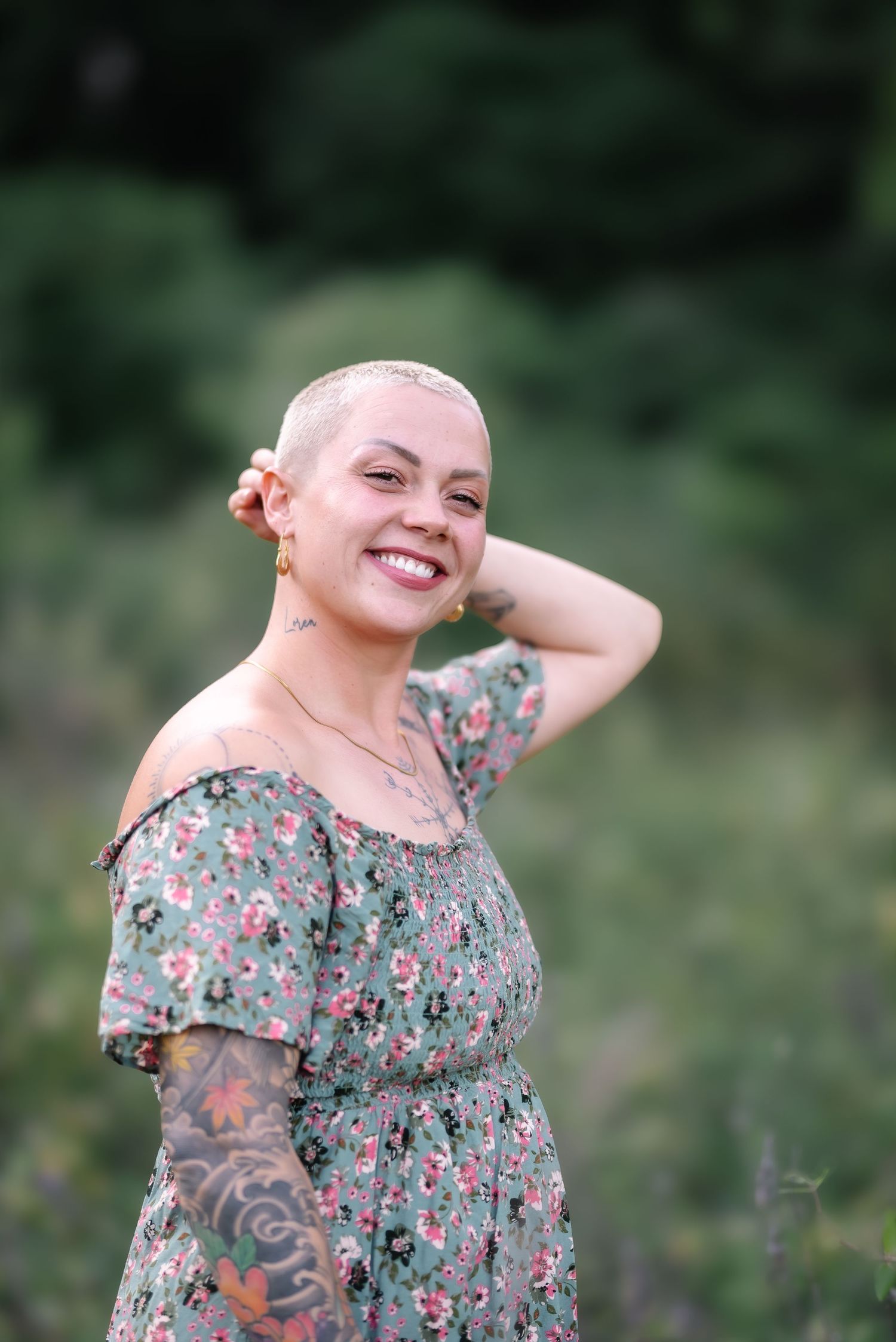 Smiling woman with shaved head, floral off-shoulder dress, and tattoos posing outdoors in a green field.