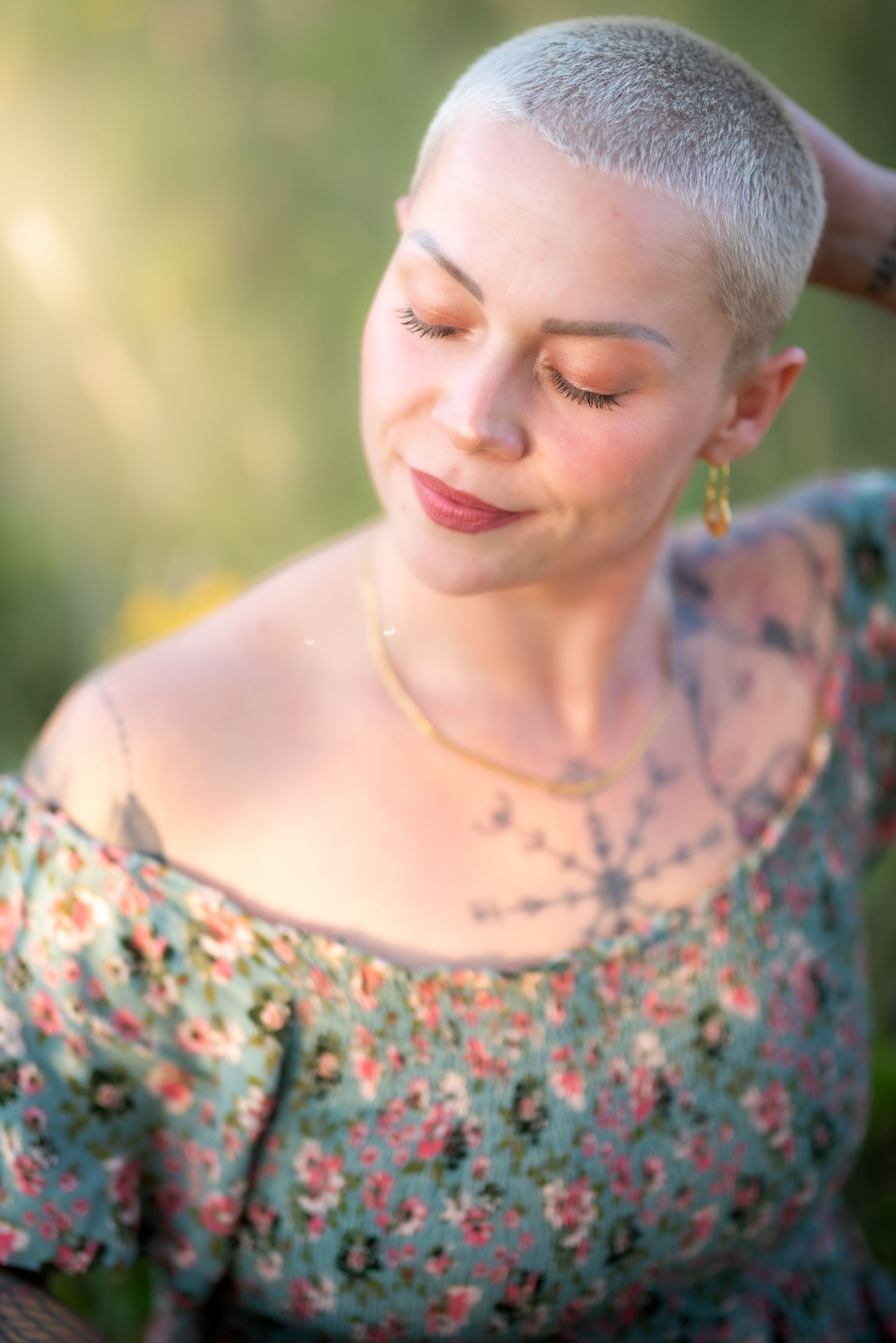 Woman with short blonde hair in floral dress, eyes closed, smiling softly in golden sunlight outdoors.