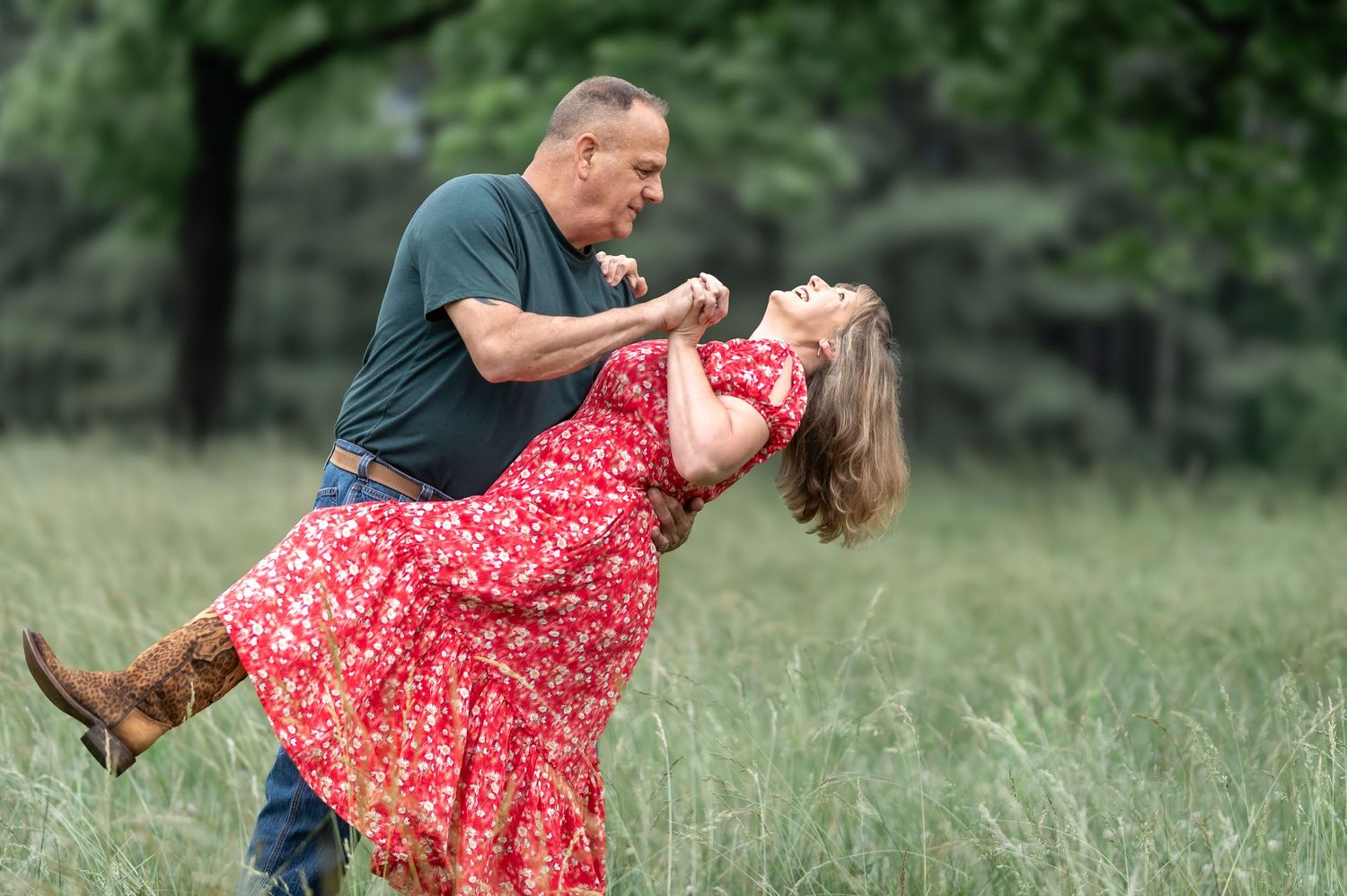 Couple dancing joyfully in a green meadow, man dipping woman in red floral dress, outdoor dance photography.