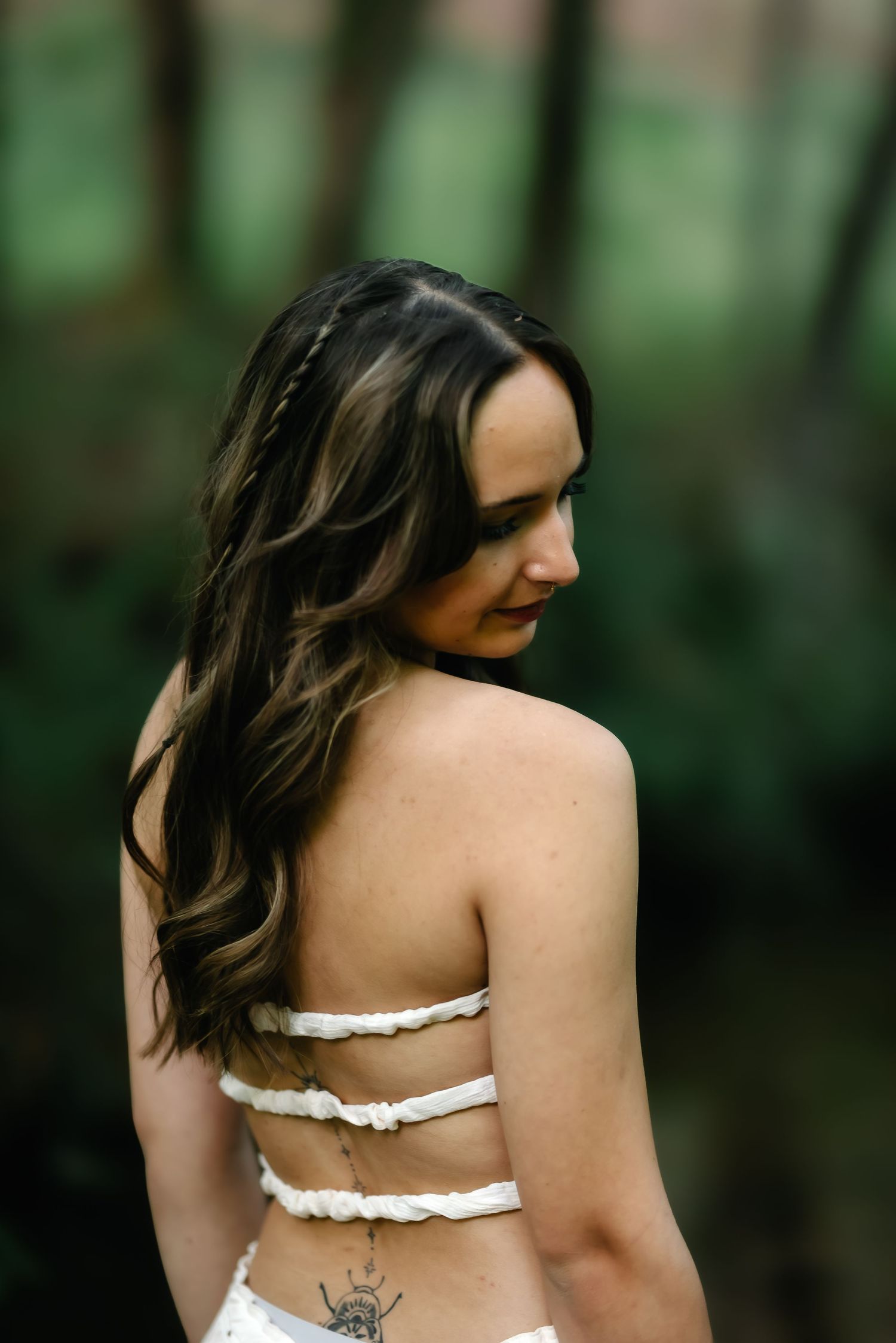 Woman with long dark hair in backless white dress with tattoo, standing in forest setting.