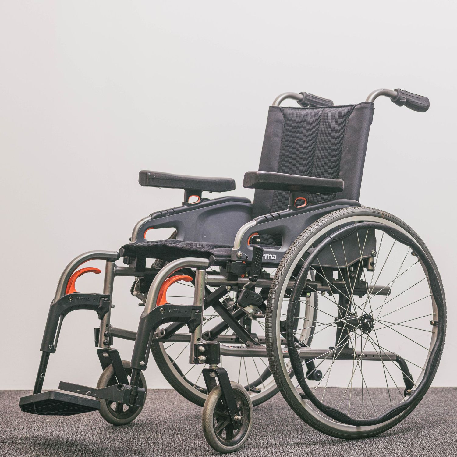 Lightweight foldable manual wheelchair with black upholstery, armrests, footrests, and large rear wheels on gray floor.
