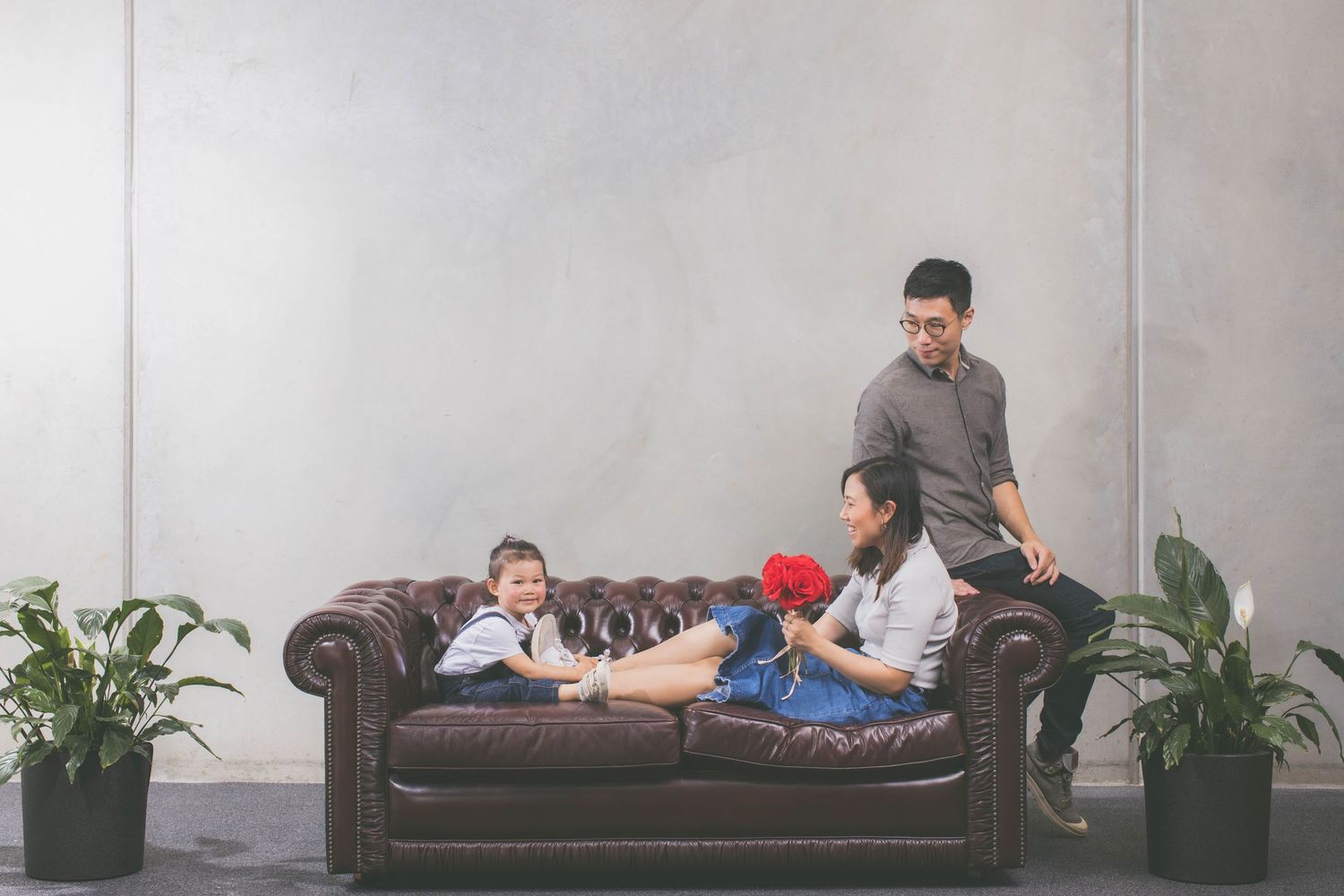 Asian family relaxing on brown leather sofa with two children, plants, and gray wall in modern living room.