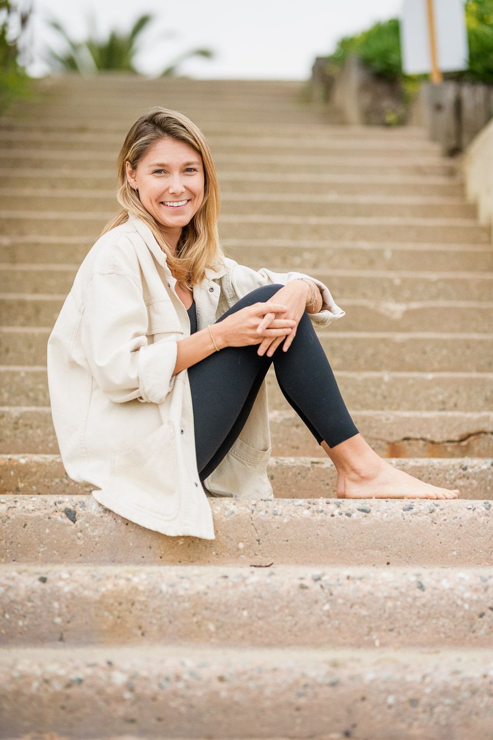 Smiling woman sitting barefoot on outdoor stone steps wearing black leggings and a white jacket.
