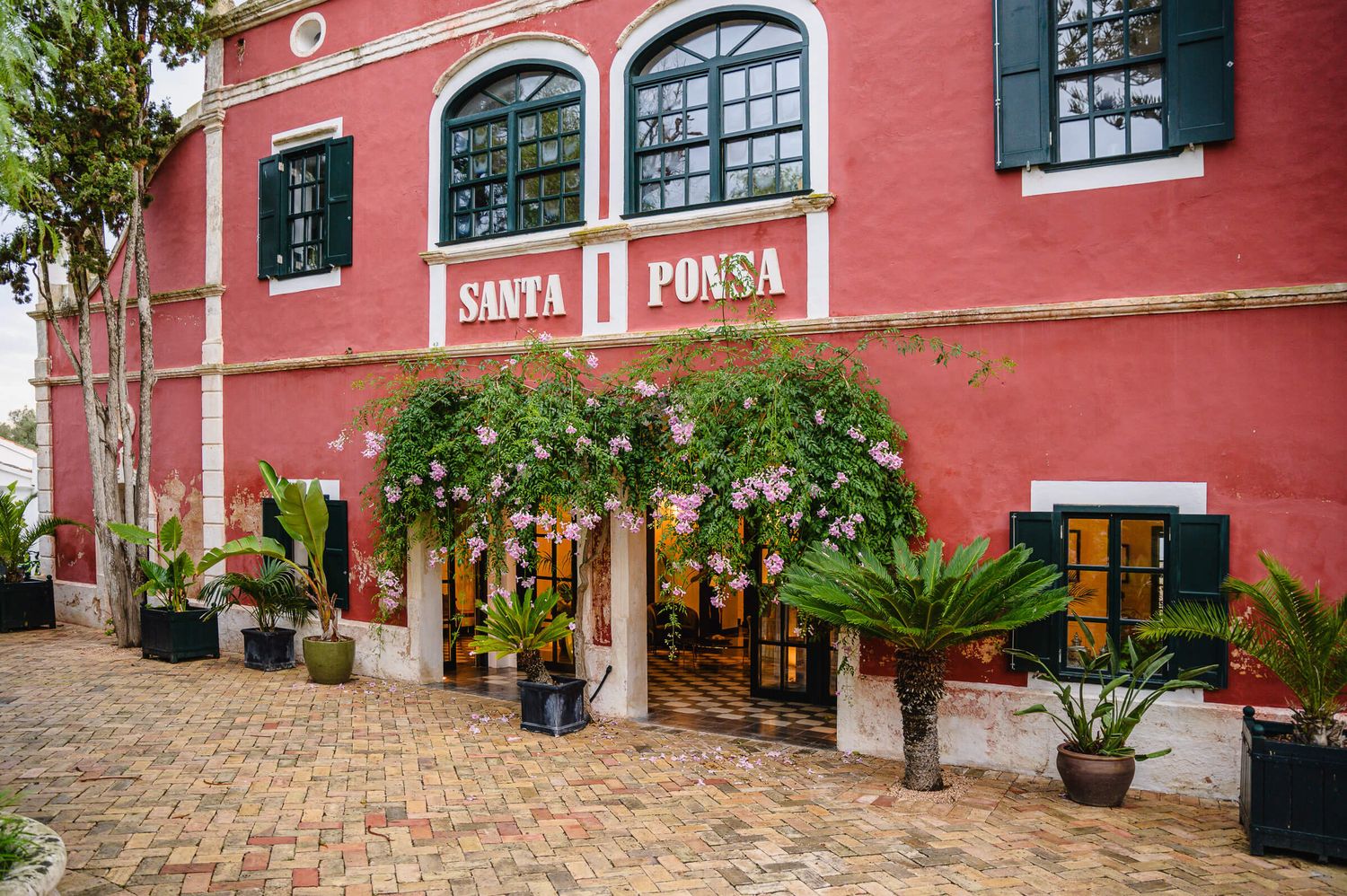 Santa Ponsa restaurant exterior with red facade, lush tropical plants, and elegant arched windows on a brick courtyard.