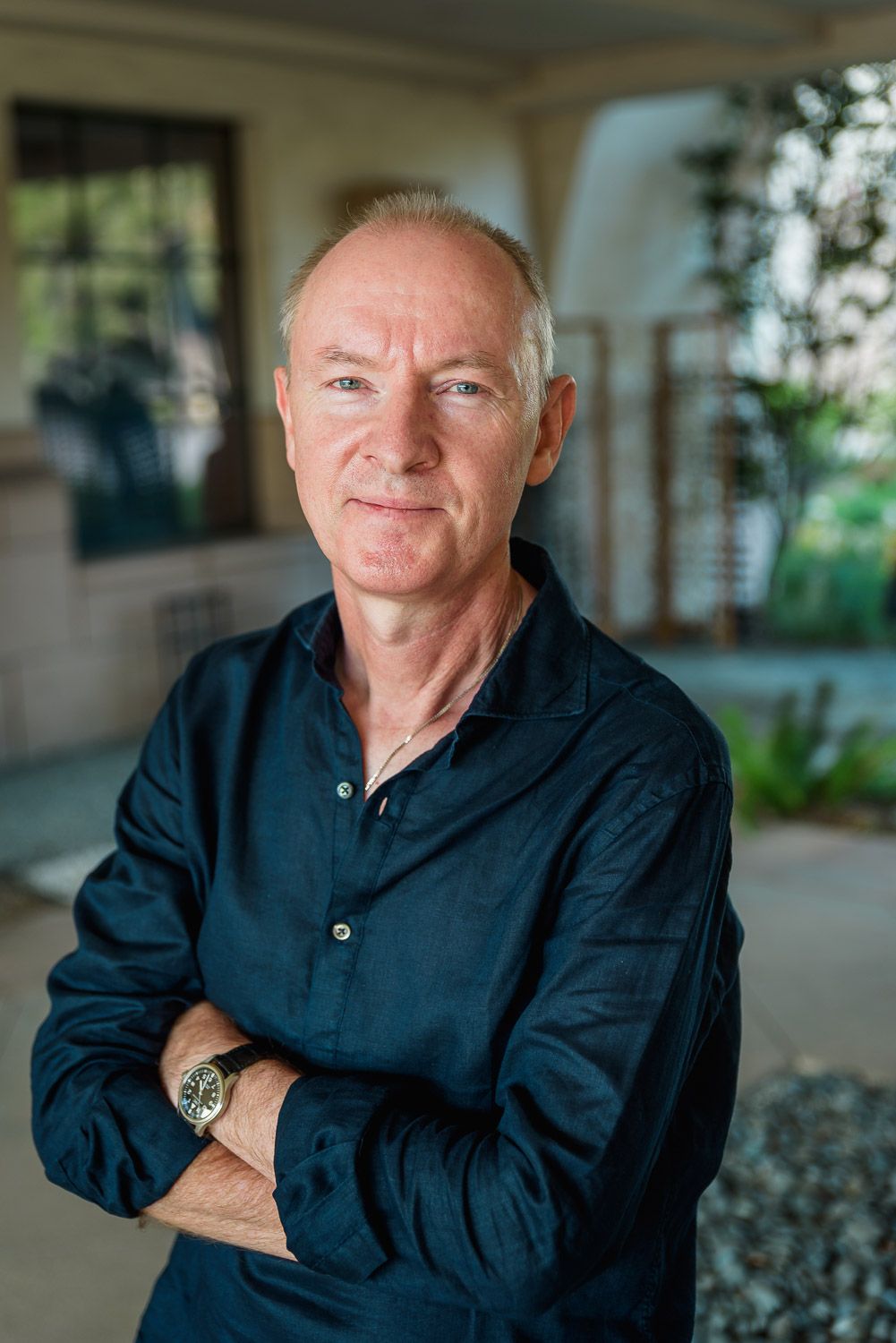 Middle-aged man with gray hair stands arms crossed outdoors, wearing a dark blue shirt and watch.