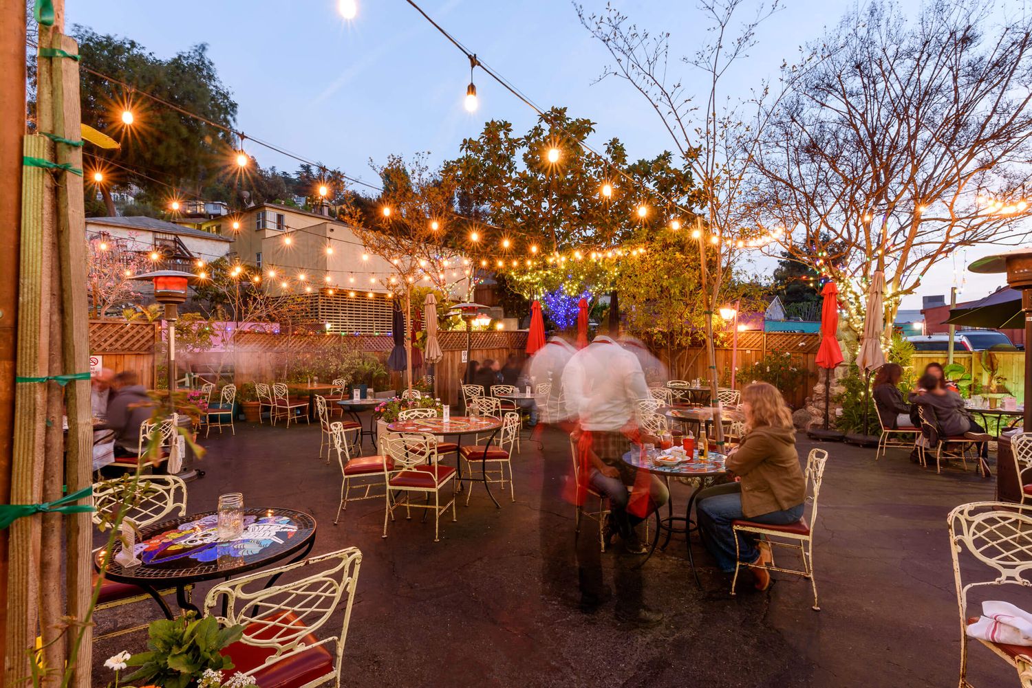 Charming outdoor patio restaurant at dusk with string lights, lush trees, and guests dining in cozy white chairs.