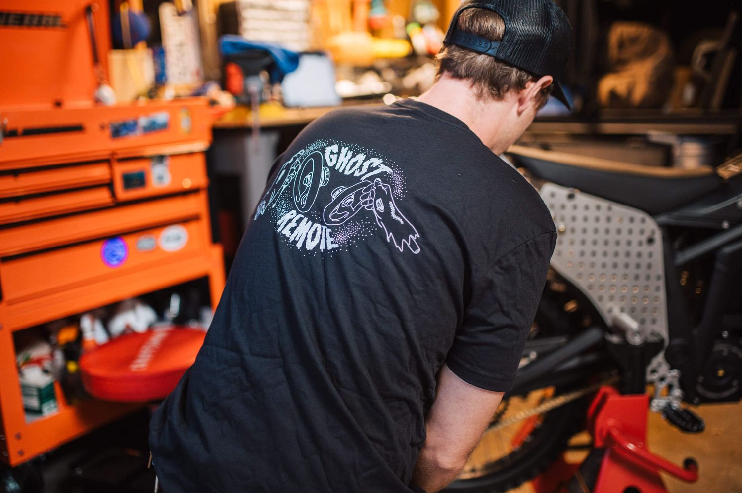 Mechanic in black graphic t-shirt with Ghost Remote logo working on a motorcycle frame near an orange tool chest.