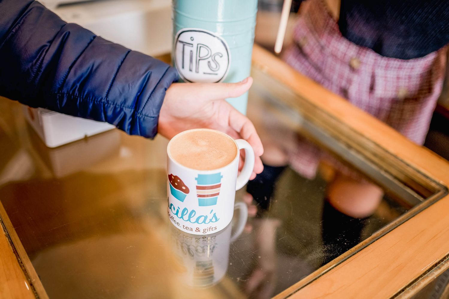 A hand placing a branded coffee mug on a glass counter near a tips jar in a cozy cafe.